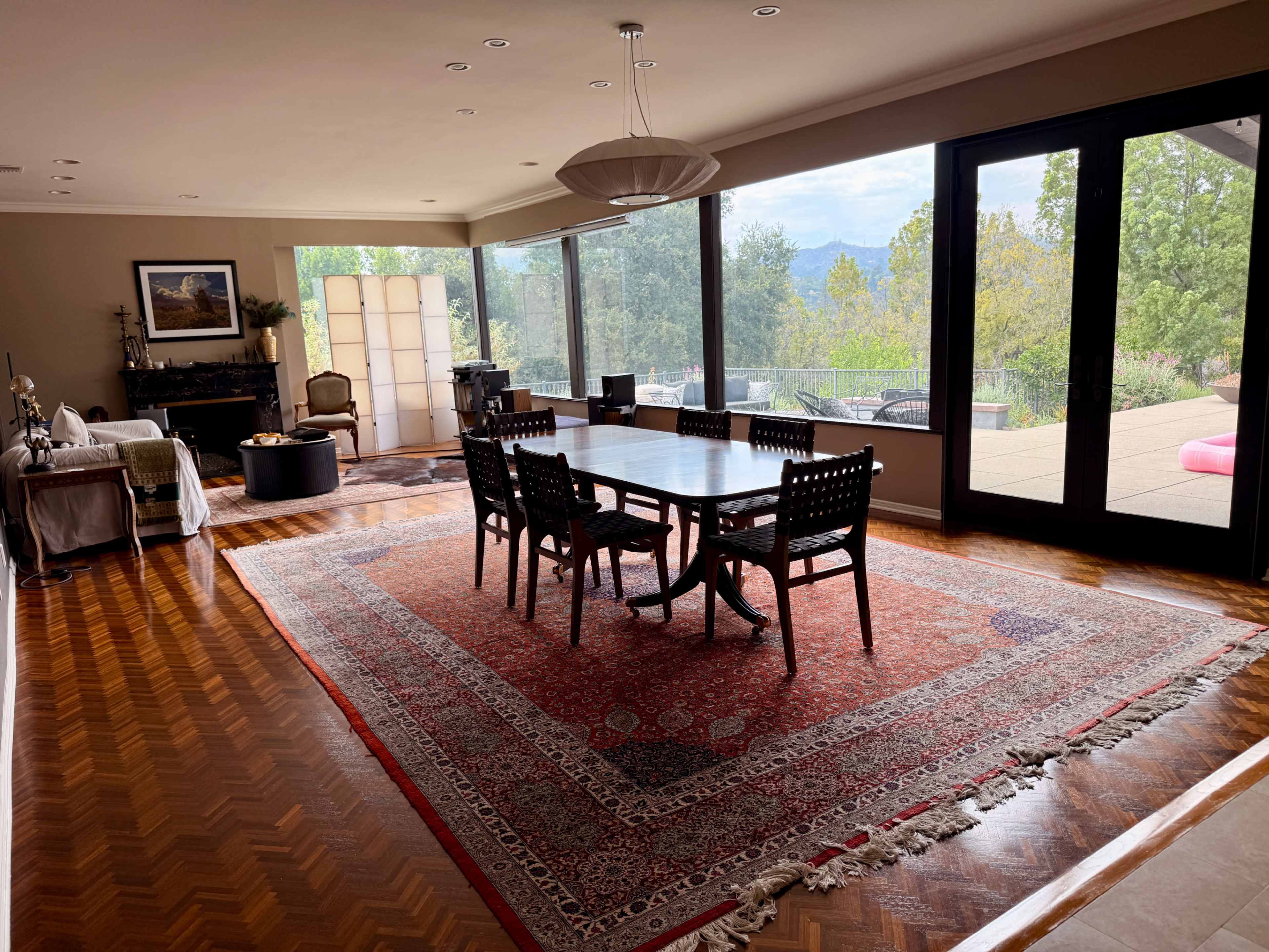 The image depicts a spacious dining area featuring a wooden table surrounded by six chairs, with large windows offering a view of the outdoors and an area rug on a hardwood floor.