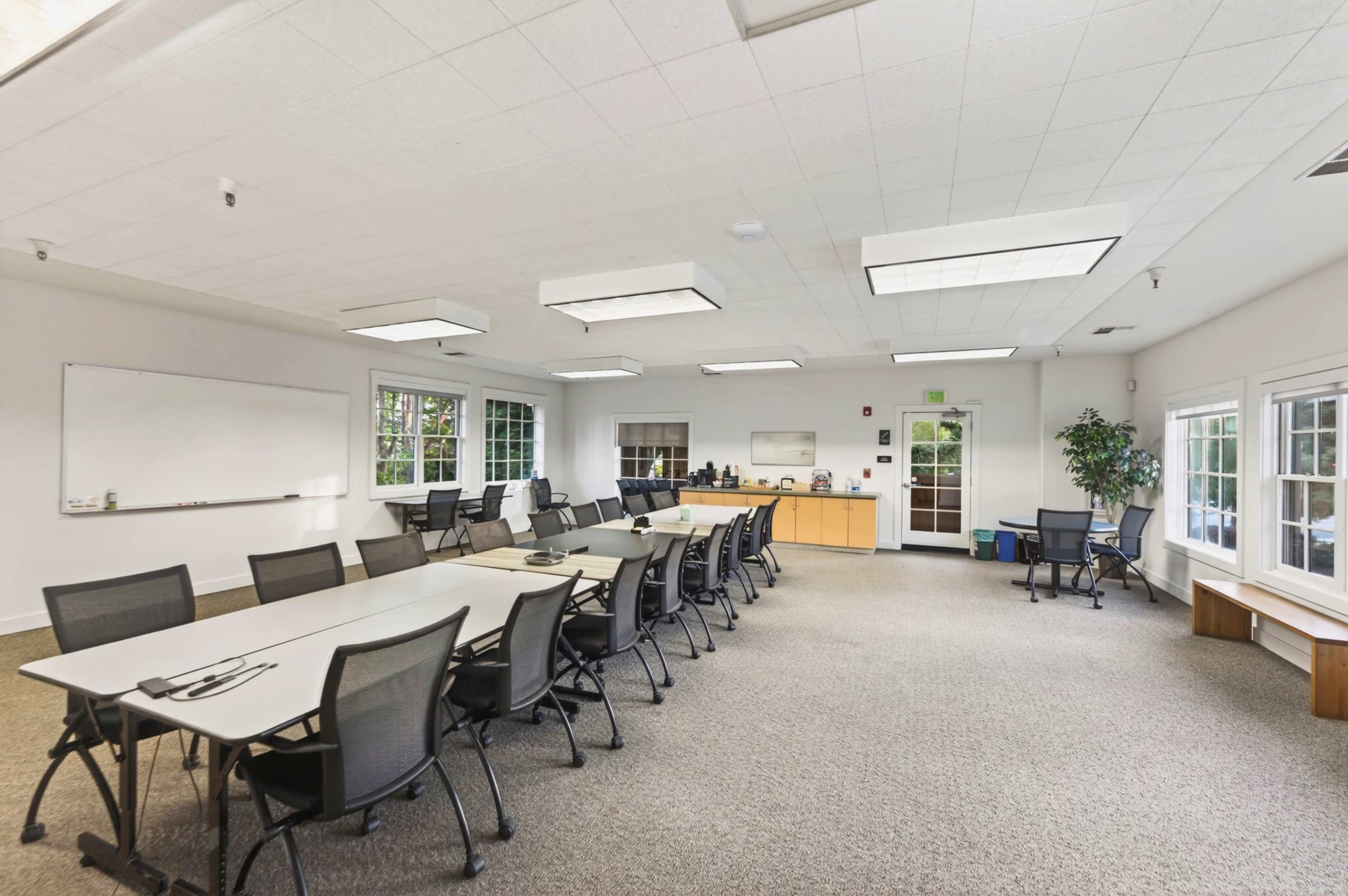 The image shows a spacious conference room with multiple long tables lined with chairs, large windows providing natural light, and a credenza against one wall.