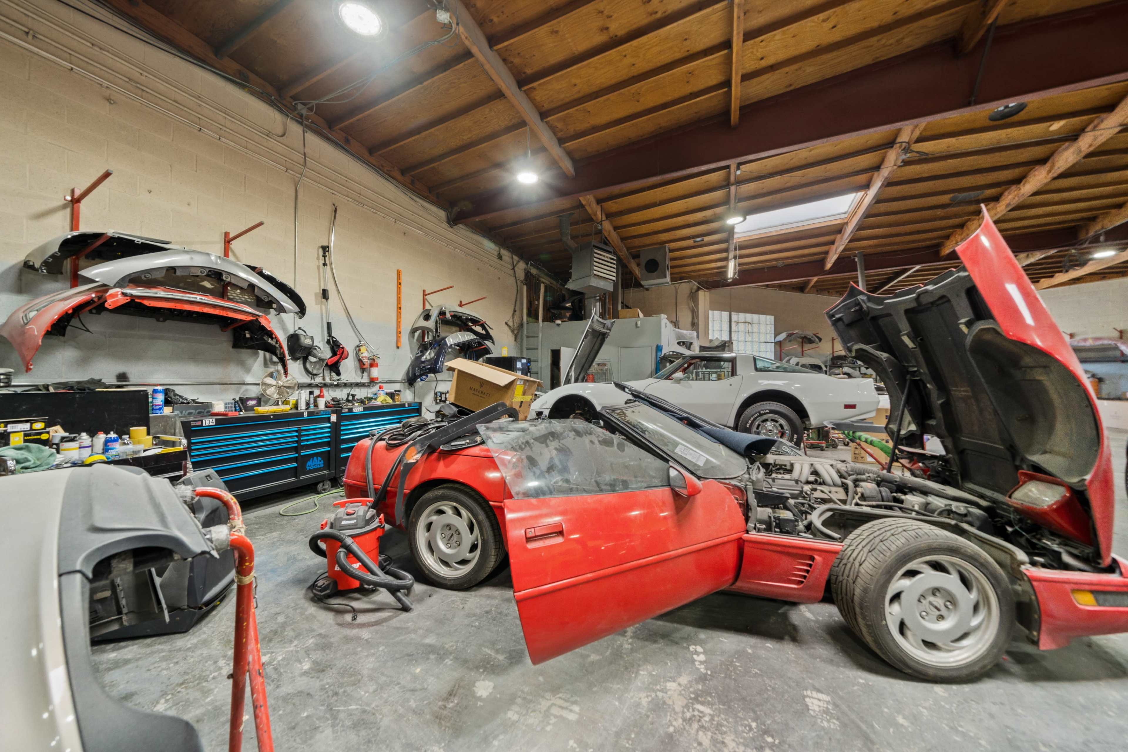 A cluttered auto repair shop contains several disassembled cars, tools, and equipment, with an open red car in the foreground.