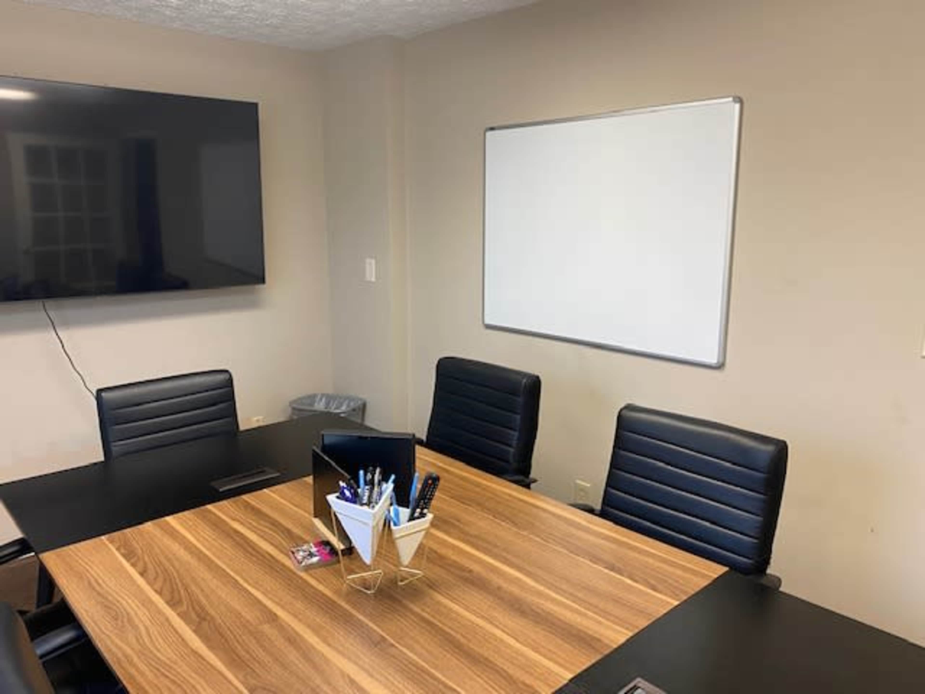 A conference room features a wooden table surrounded by four black chairs, a wall-mounted television, and a whiteboard.