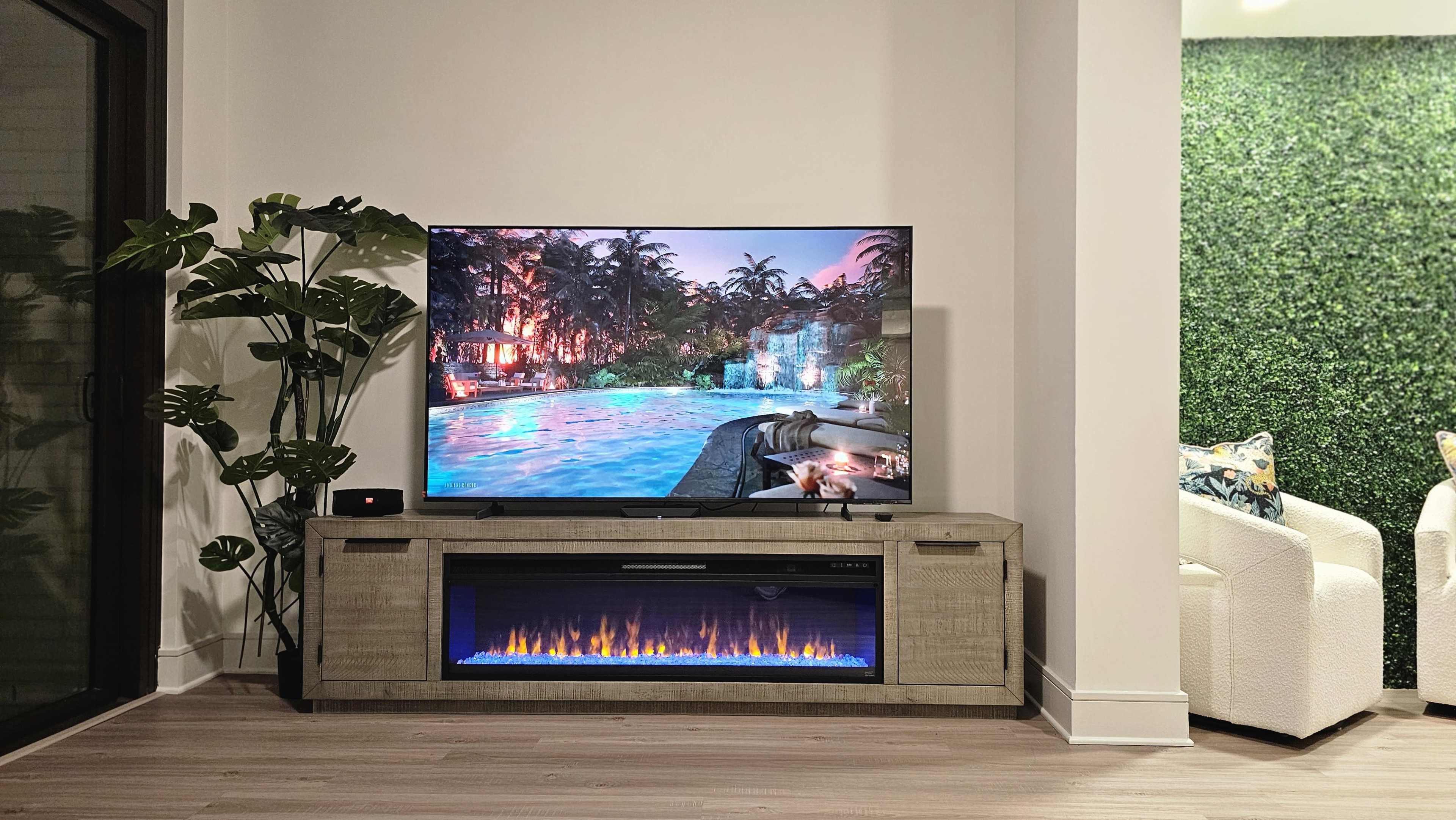 A large television displaying a poolside scene is mounted above a wooden media console featuring an electric fireplace, surrounded by indoor plants and a green wall.