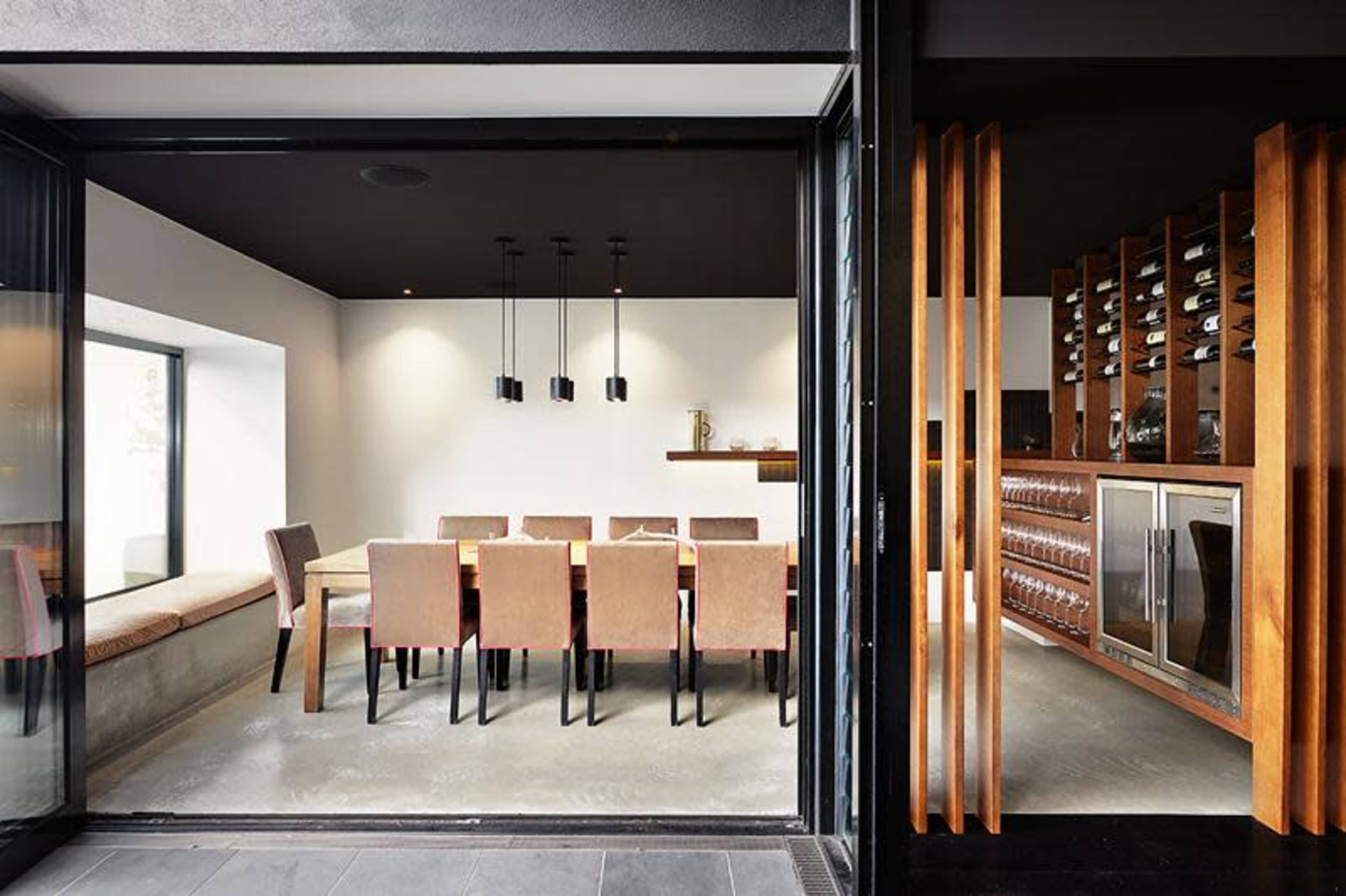 The image shows a modern dining area with a long table surrounded by chairs, adjacent to a wine storage display.