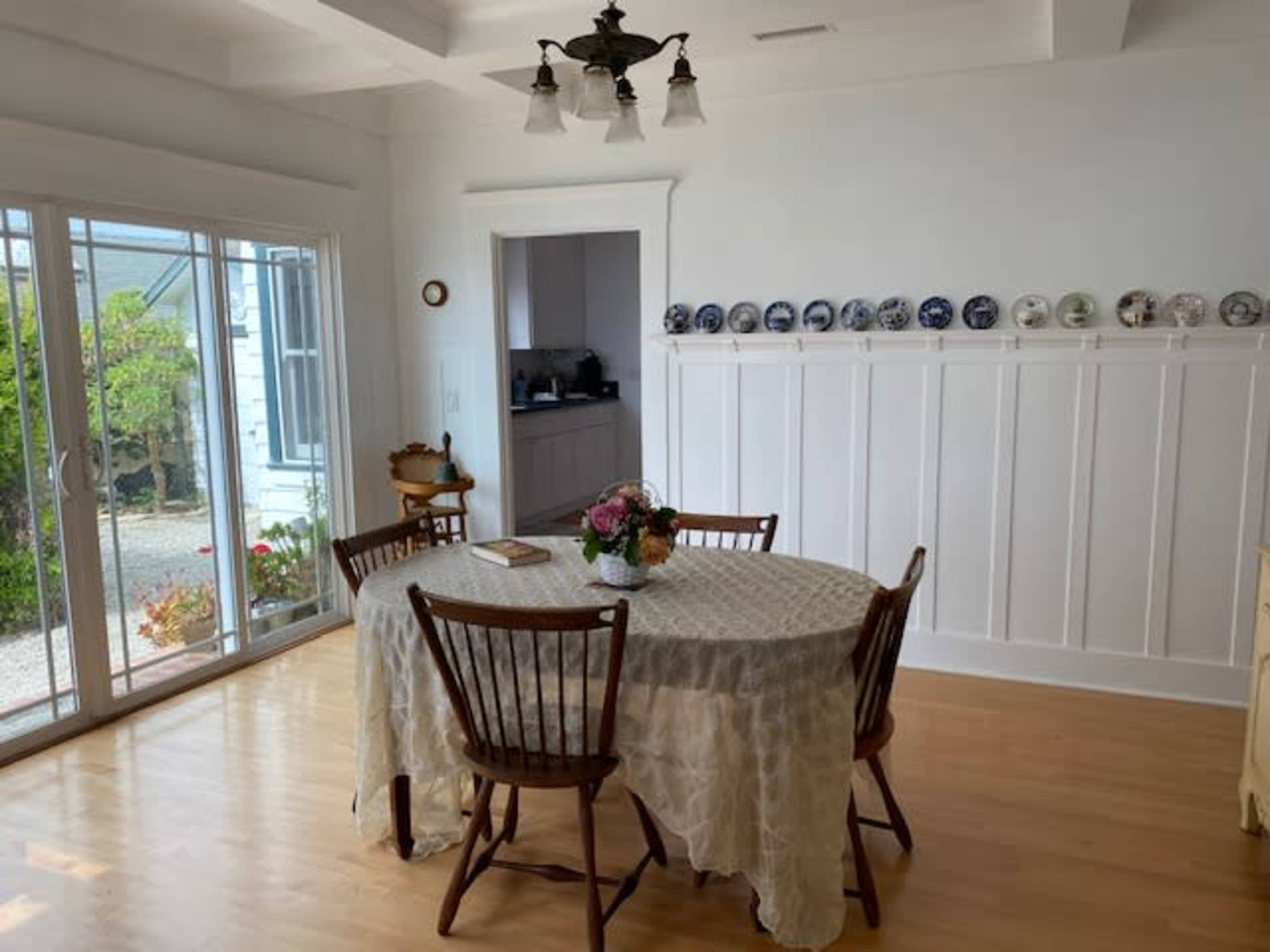 A dining area features a round table surrounded by wooden chairs, with a bouquet on the table and decorative plates mounted on the wall.