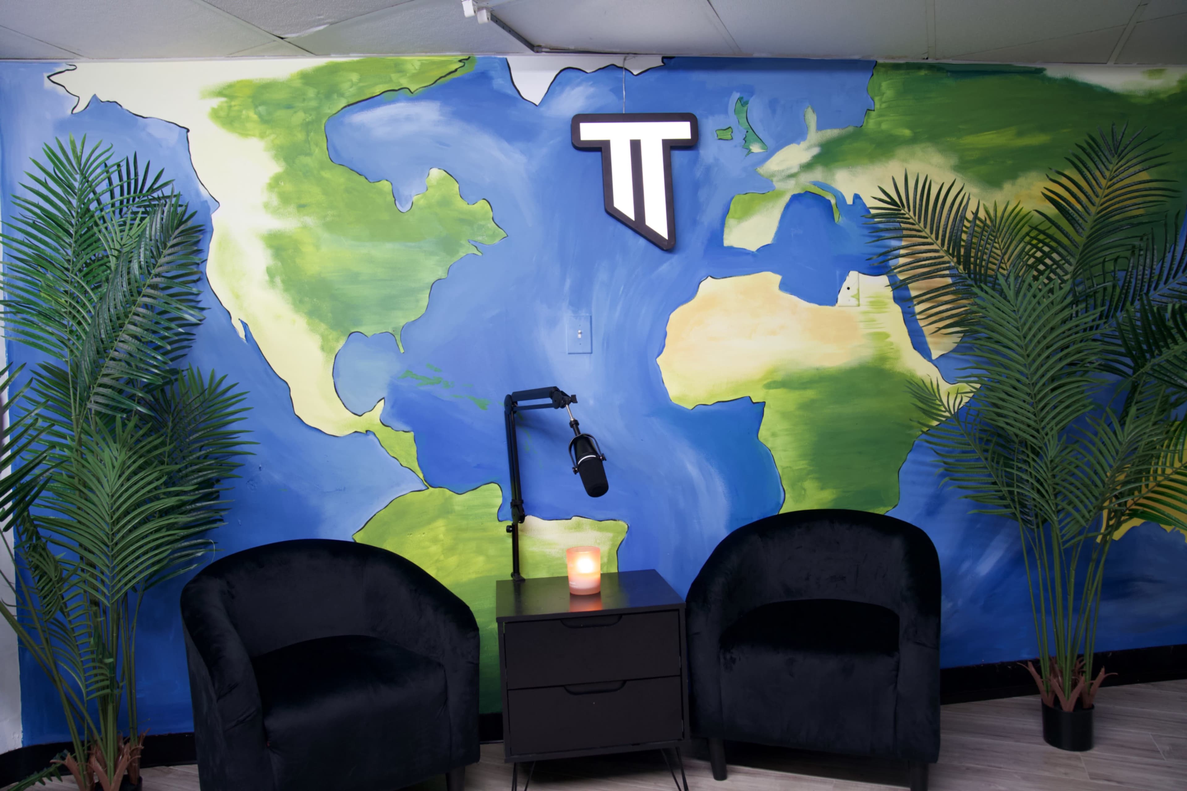 The image shows a cozy seating area with two black chairs, a small table with a candle, and a large mural of a world map on the wall, accompanied by potted plants.