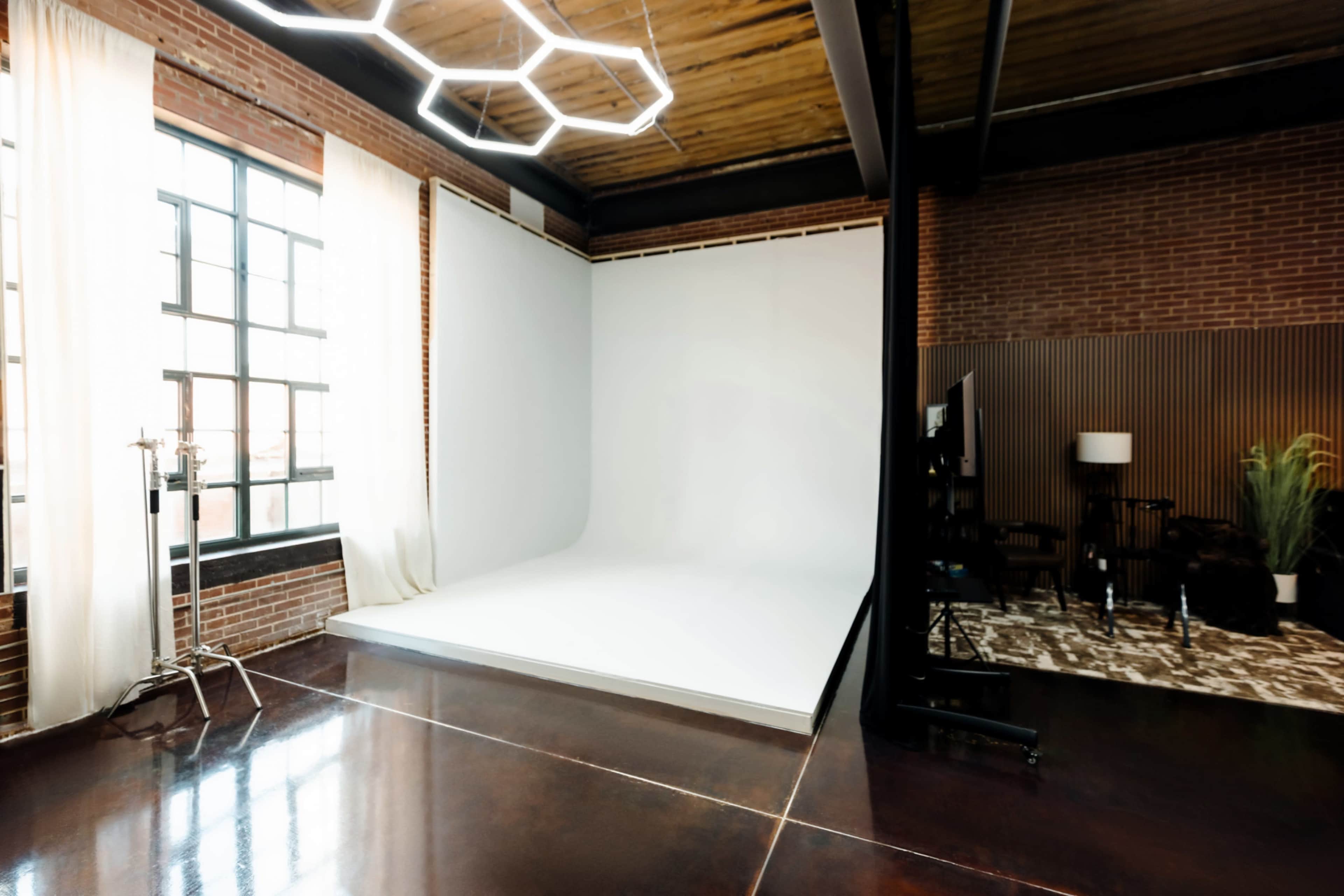 The image shows a spacious photo studio with a white backdrop, large windows, and a modern lighting fixture.