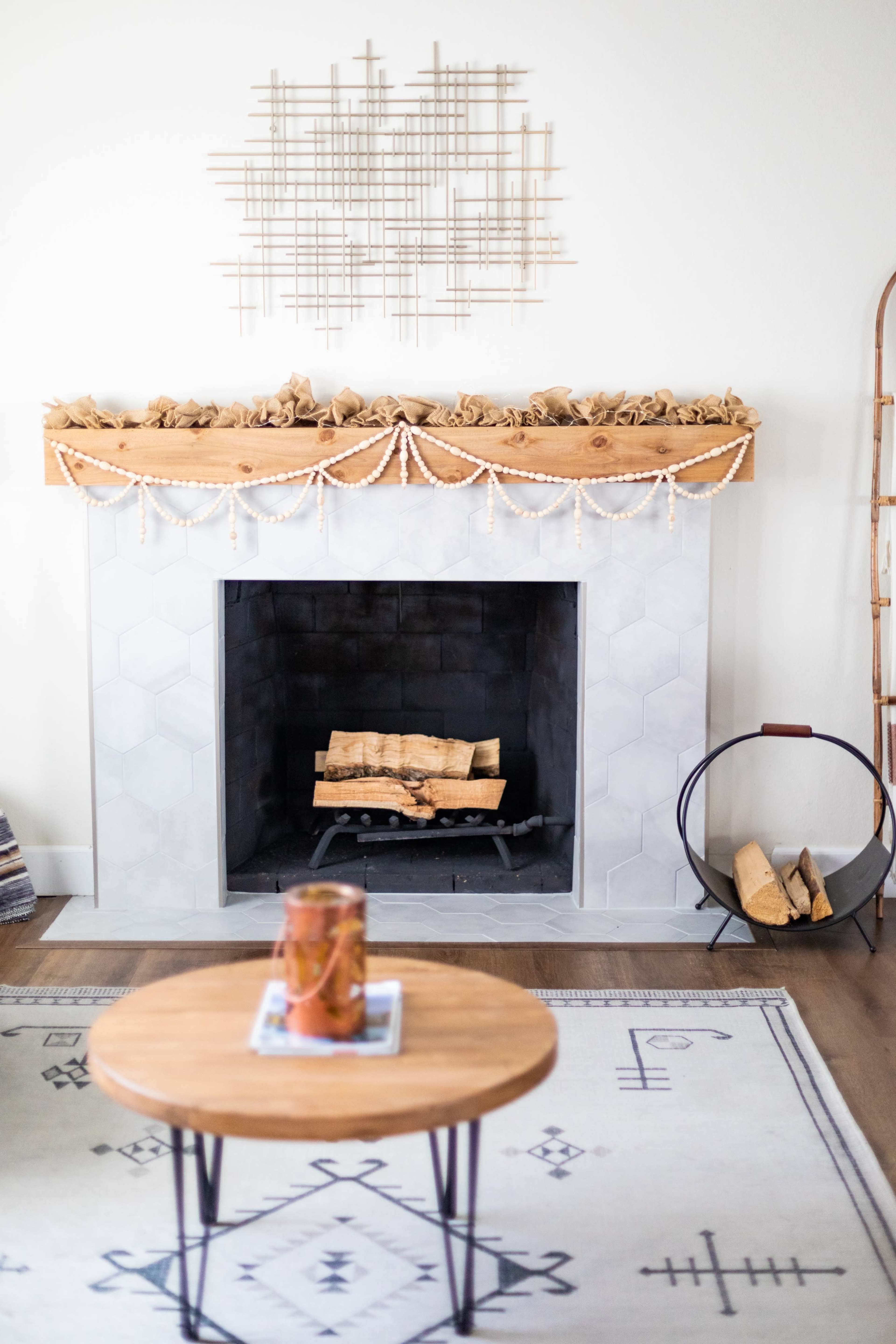 The image shows a modern fireplace with a gray hexagonal tiled surround, a wooden mantel decorated with brown paper, and a circular stand holding logs beside a round wooden table on a patterned rug.