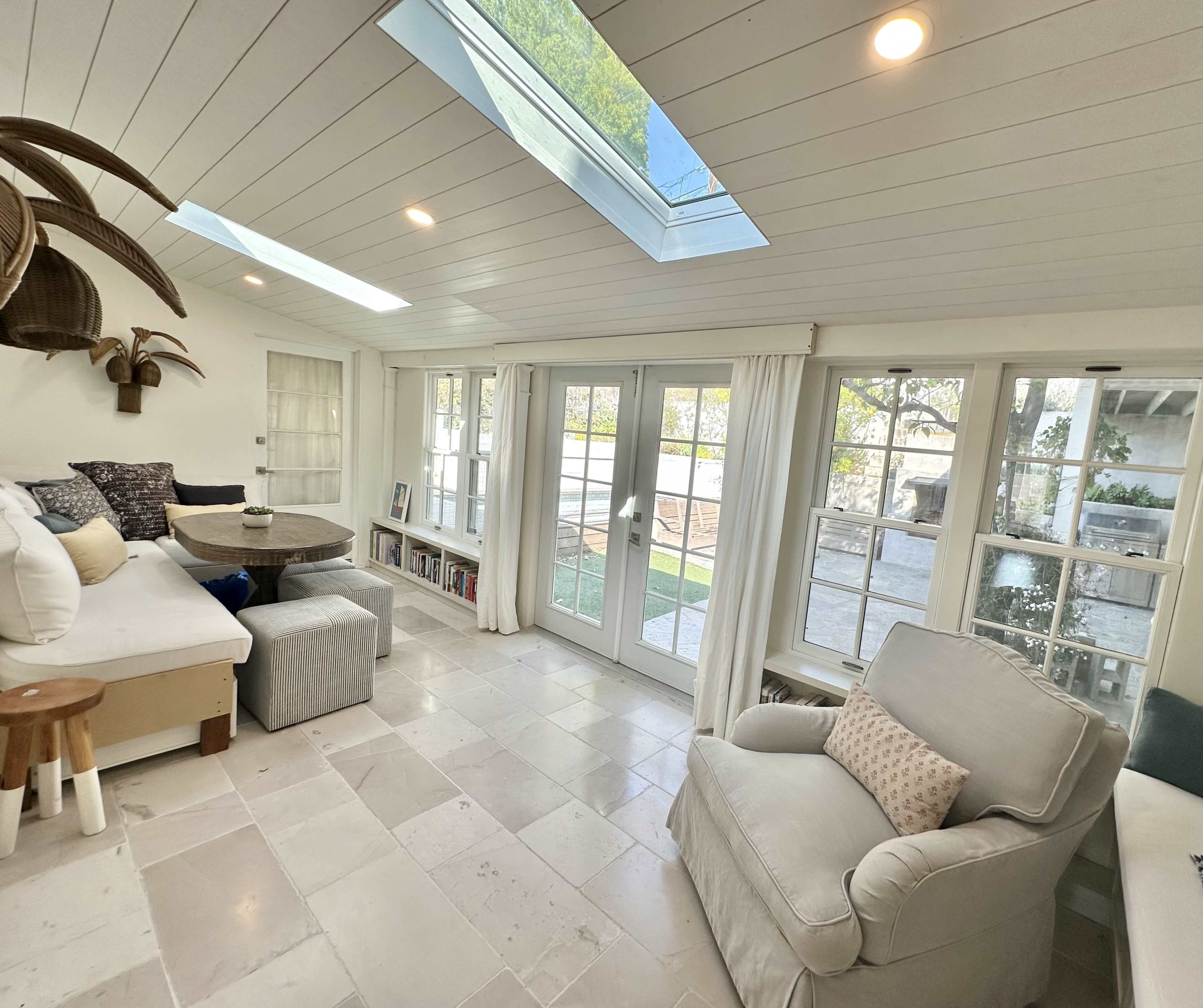 The image shows a bright, airy living room with large windows, a skylight, and a mix of comfortable seating arrangements.