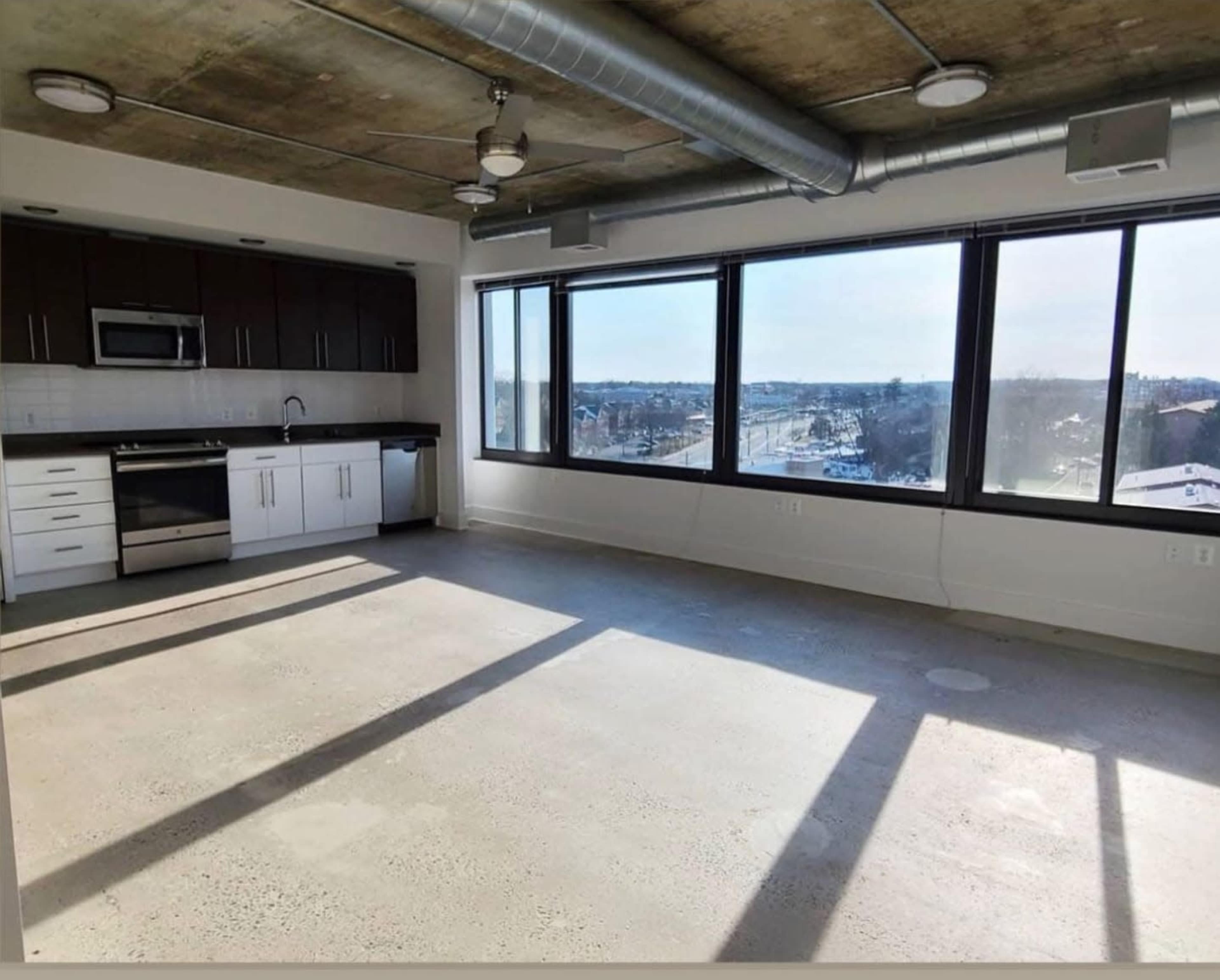An empty modern apartment with large windows, concrete flooring, and a kitchen area featuring dark cabinetry and stainless steel appliances.