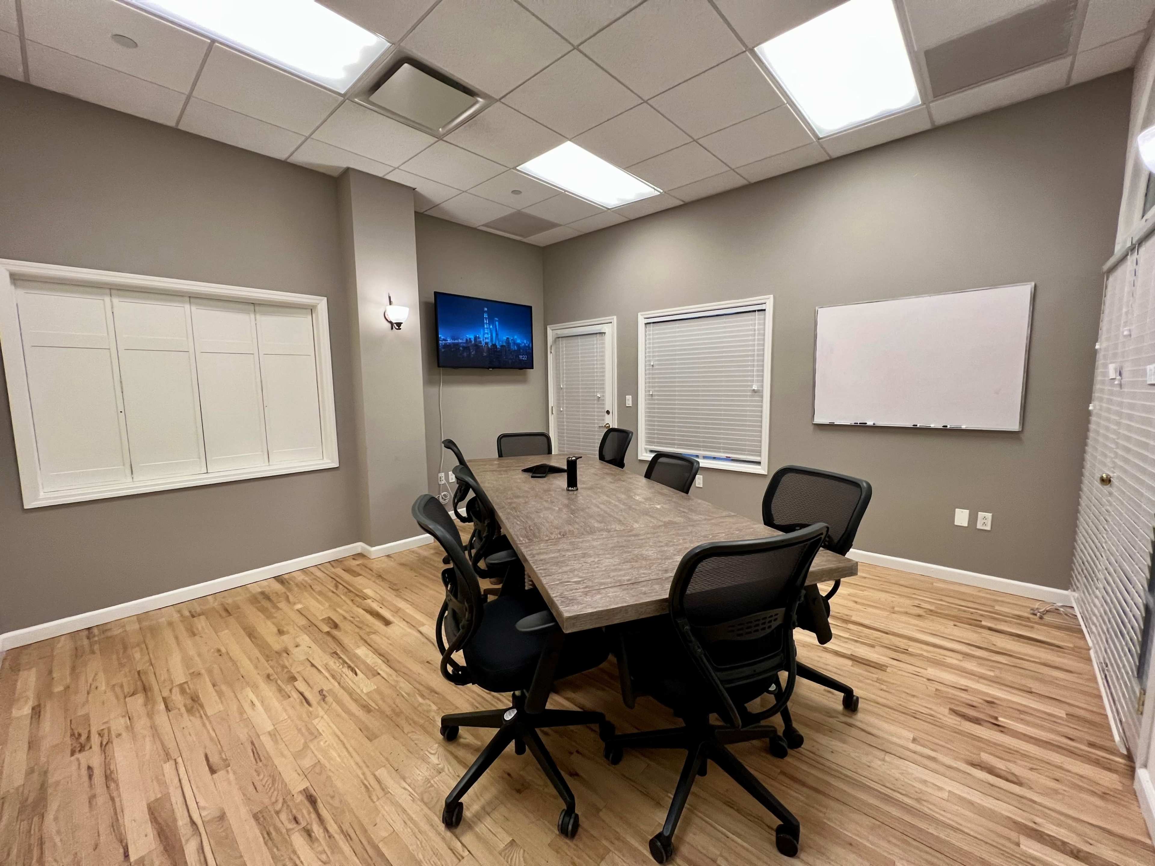A conference room features a large wooden table surrounded by black rolling chairs, with a television mounted on the wall and a whiteboard visible.
