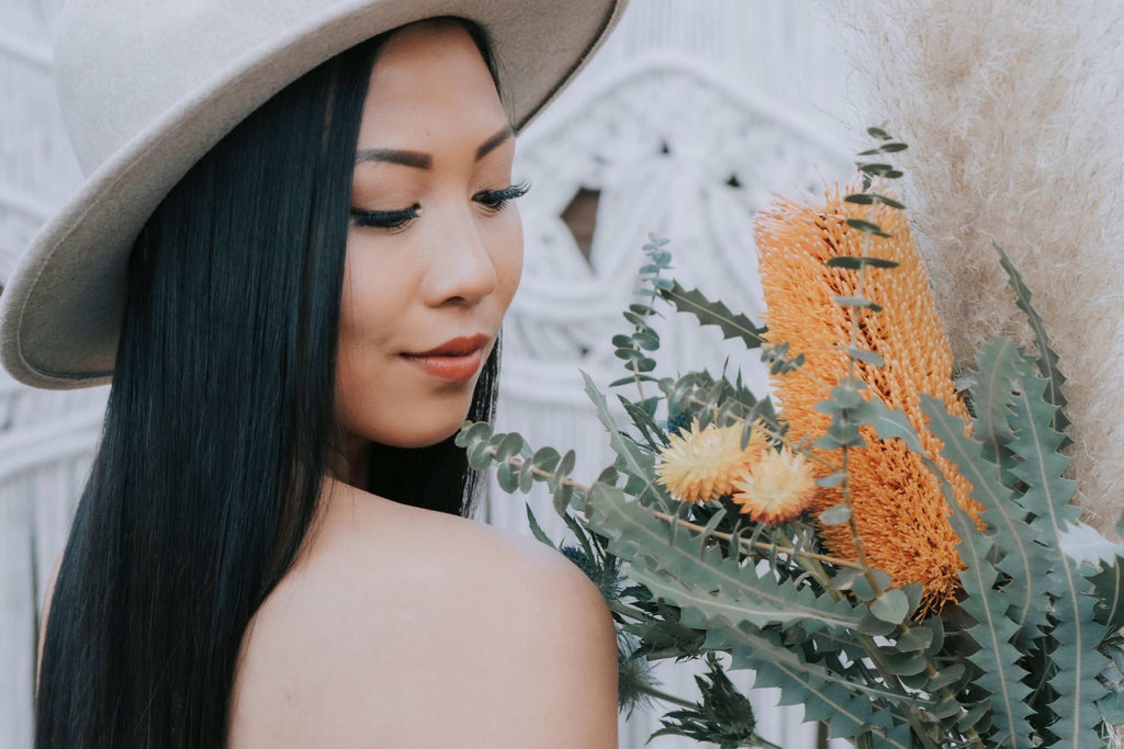 A woman with long black hair wears a wide-brimmed hat and holds a bouquet of mixed flowers and foliage.