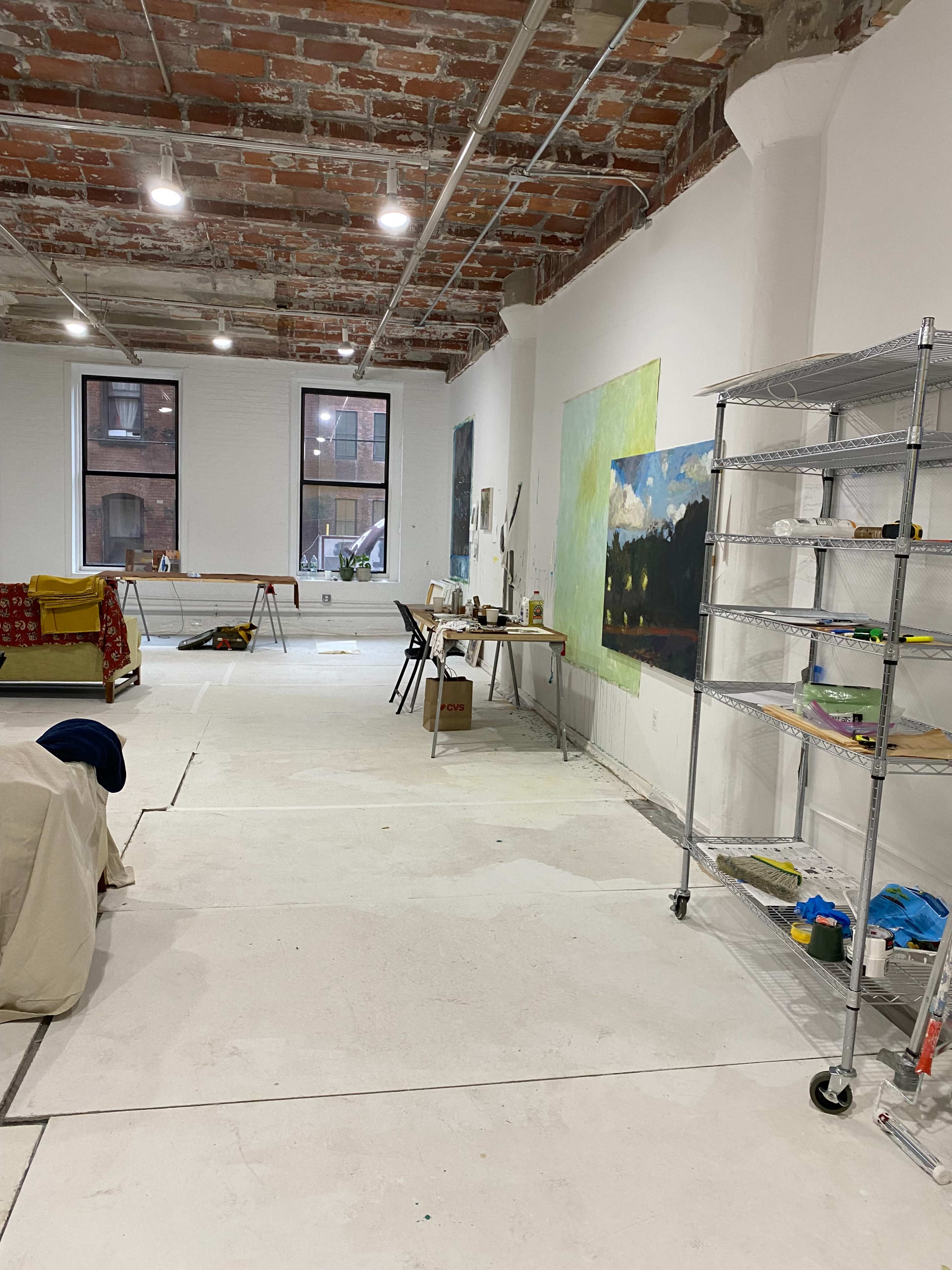 A spacious artist studio with exposed brick walls, large windows, and various painting supplies scattered around.