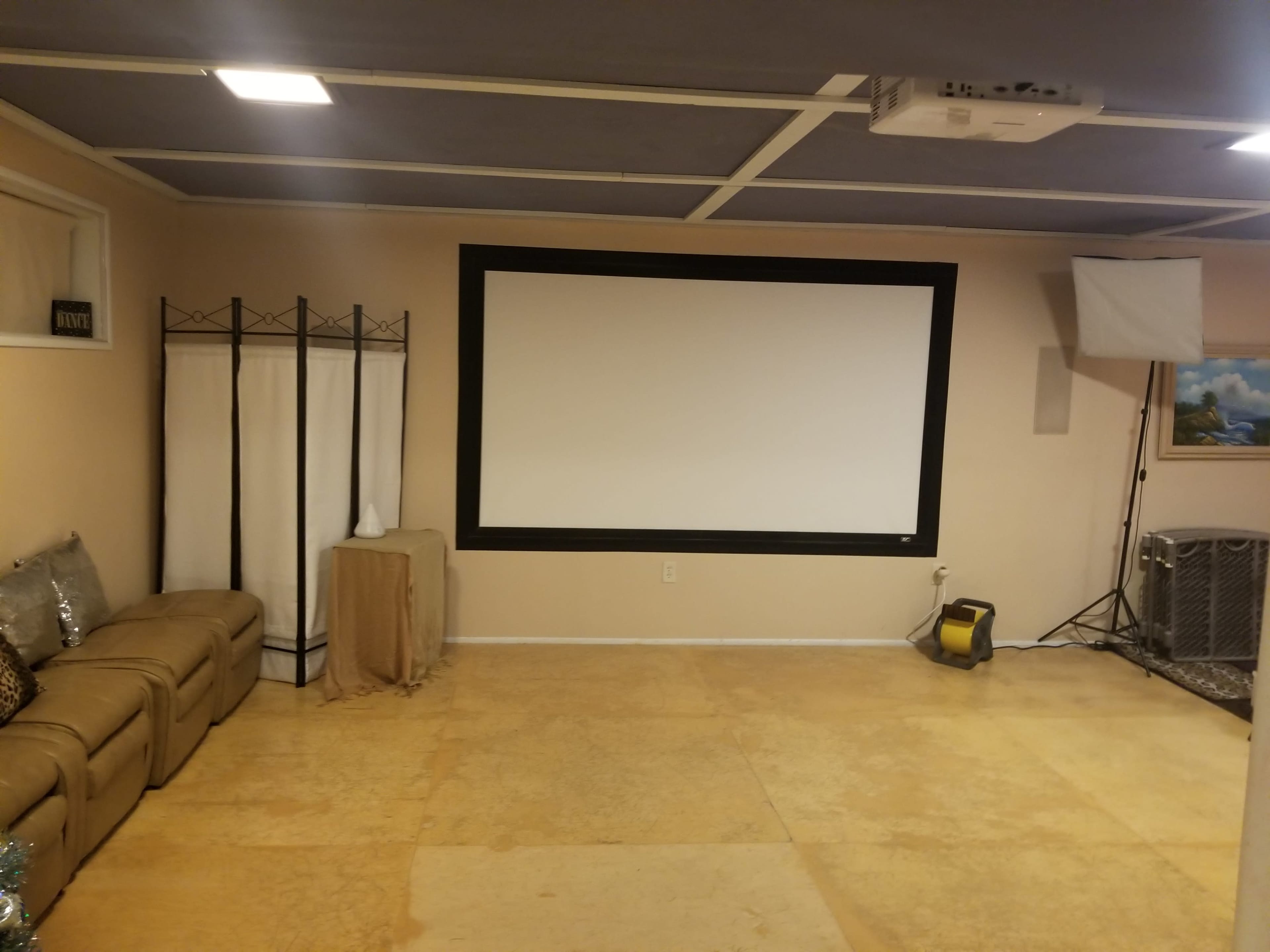 An empty room with a large projection screen on one wall, a sofa along another wall, and lighting equipment in the corner.