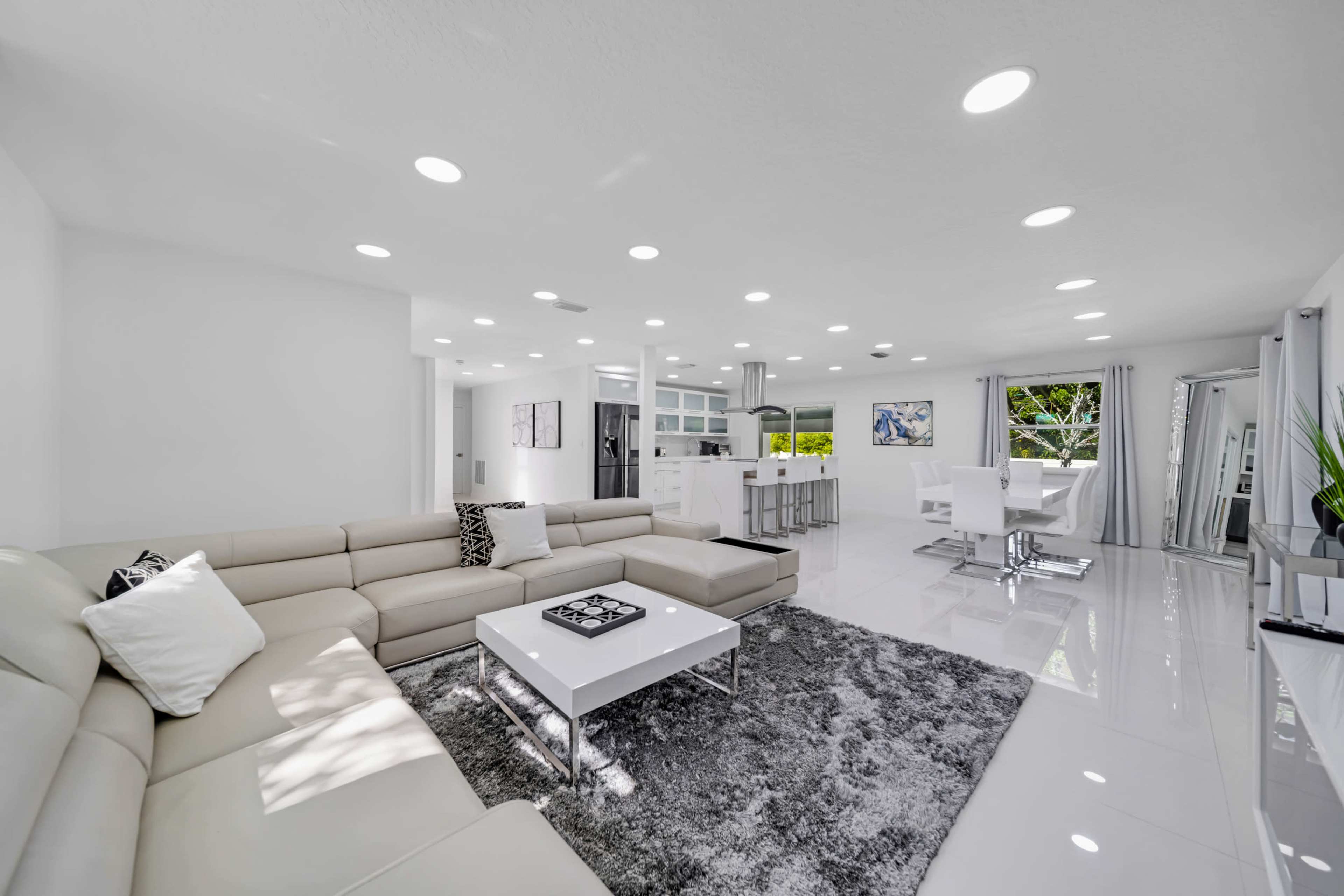 A bright, modern living room features a white sectional sofa, a coffee table, and an adjoining dining area with white furniture and large windows.