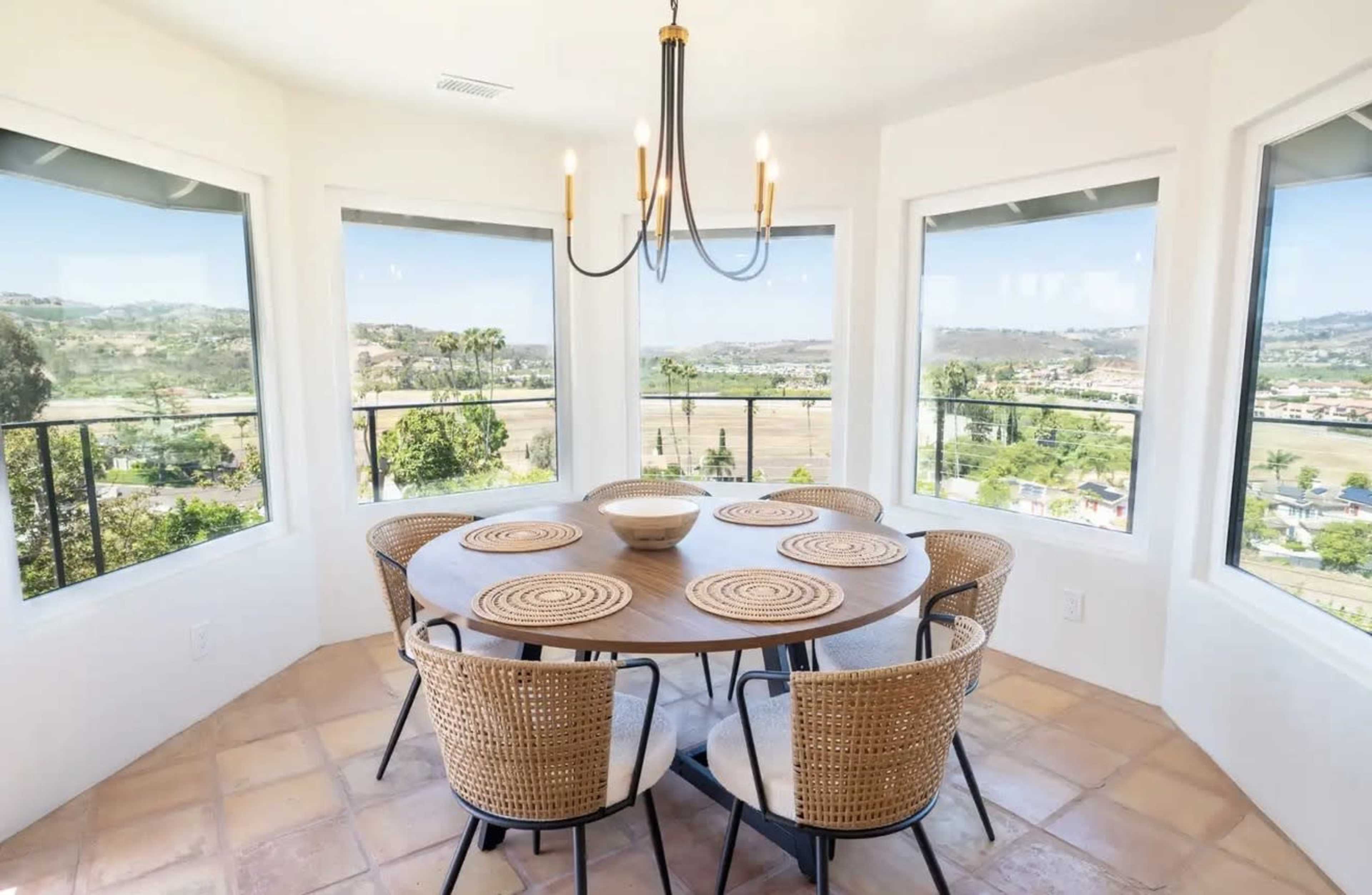 The image shows a circular dining table with woven chairs set in a bright room featuring large windows that provide a view of the surrounding landscape.