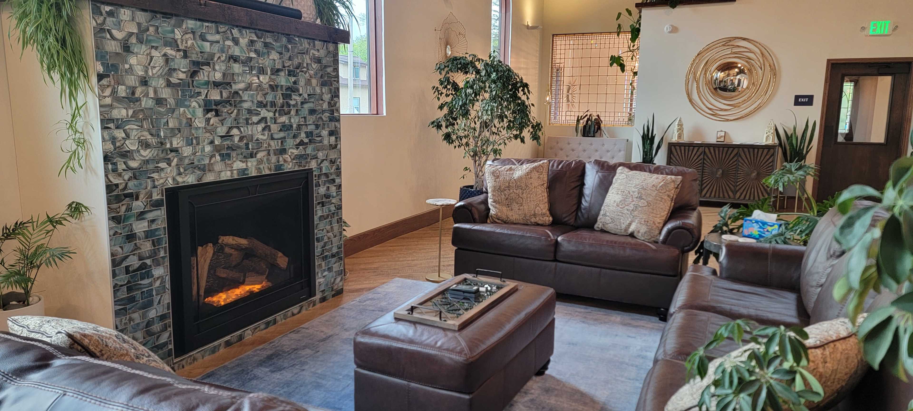 The image depicts a cozy living area featuring a stone fireplace, two brown leather sofas, a coffee table, and various indoor plants.