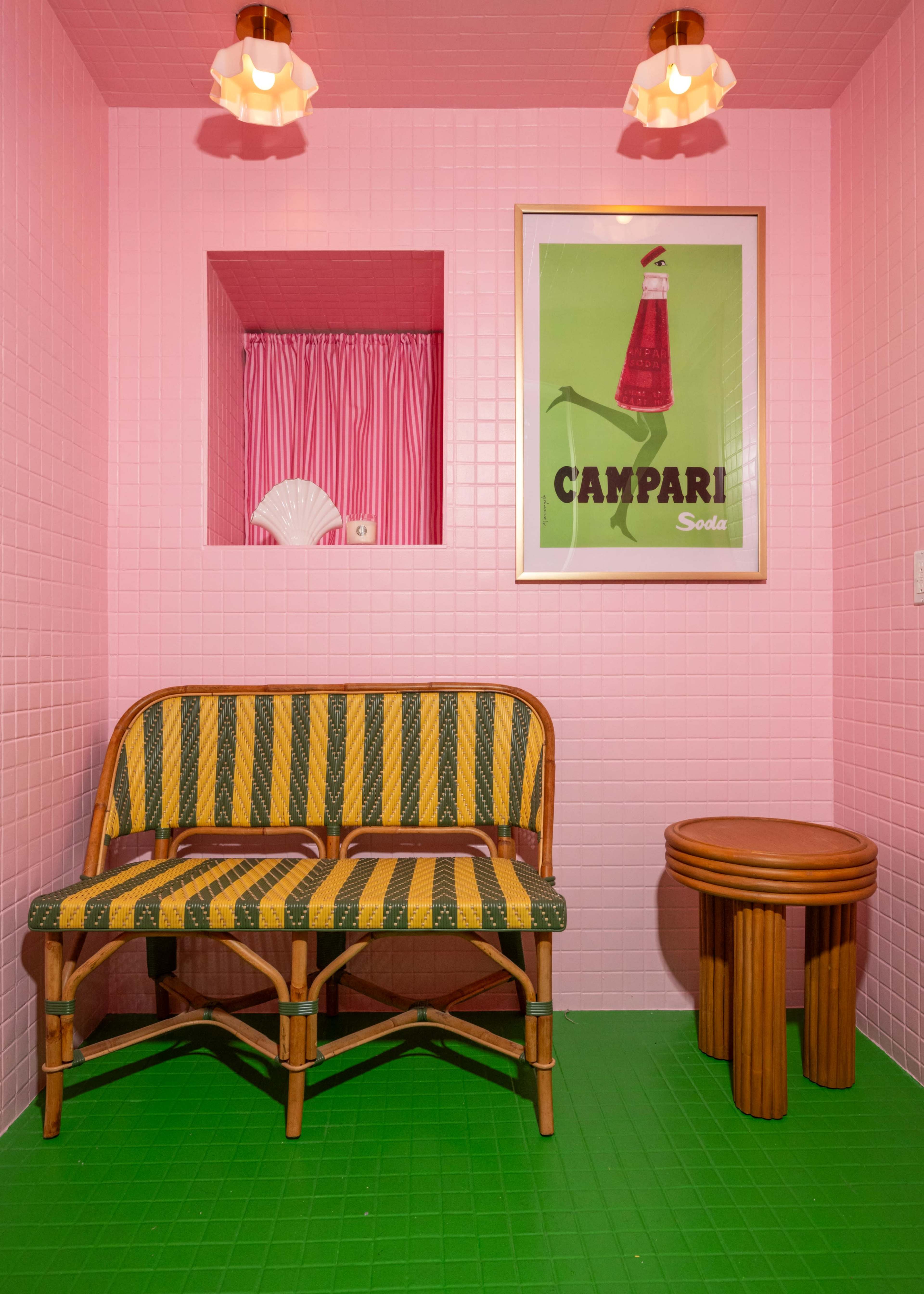 The image shows a small, brightly colored room with pink tiled walls, featuring a green floor and a vintage-style bench along with a round side table, and a framed Campari poster on the wall.