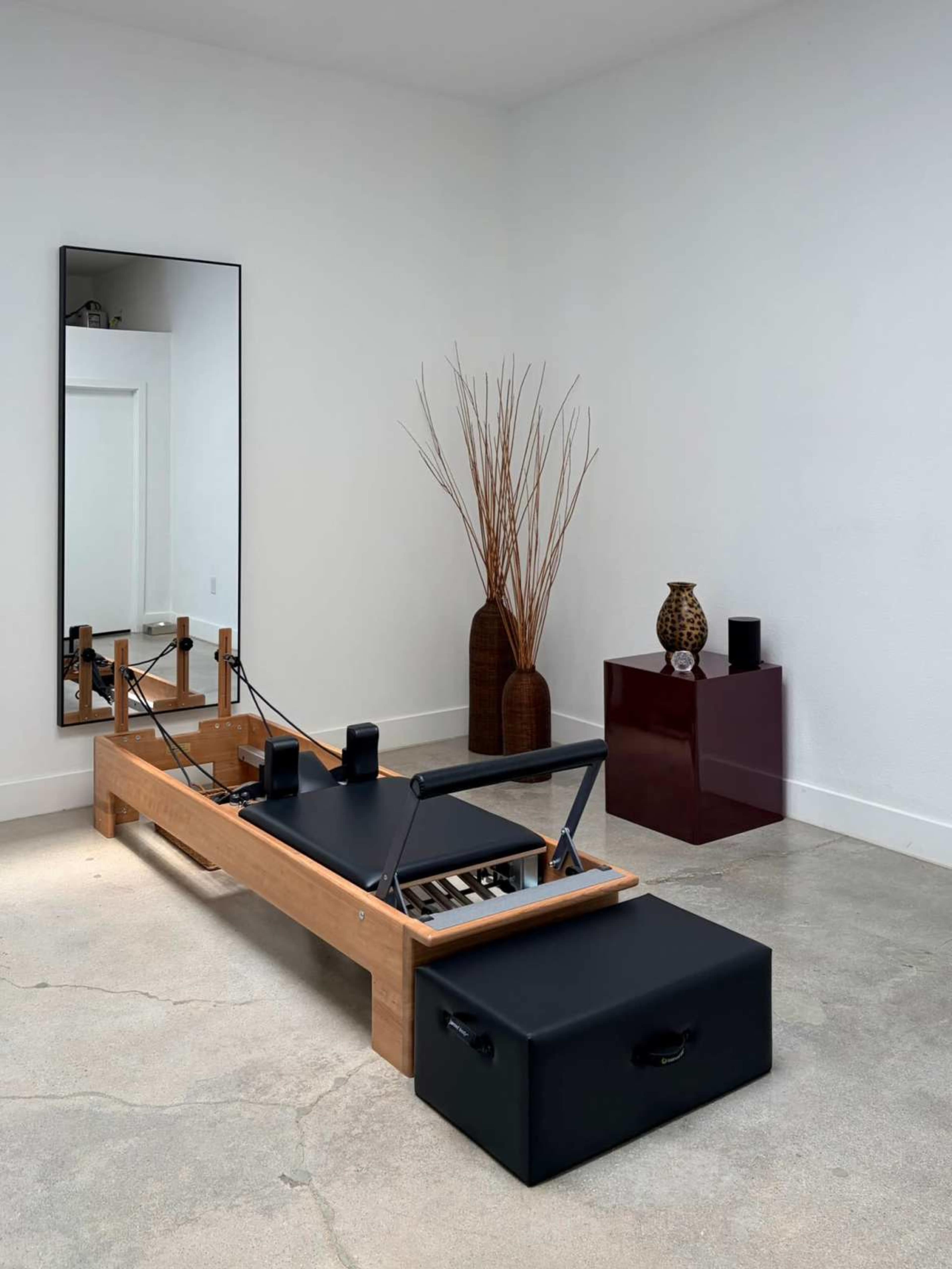 The image shows a minimalistic workout space featuring a wooden Pilates reformer, a large mirror, a decorative vase with dried plants, and a small, glossy cube table.