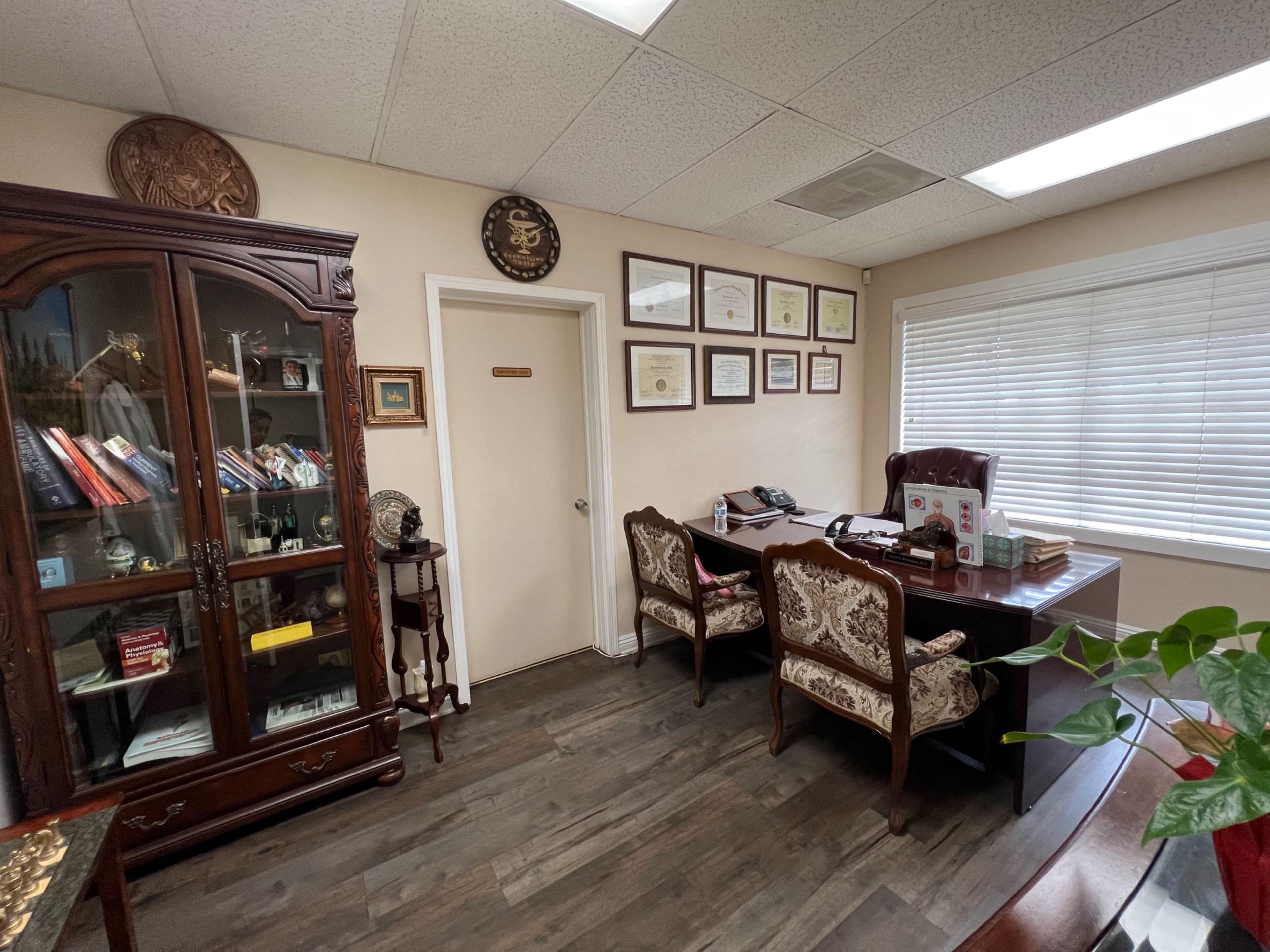 An office with a desk, two chairs, a glass display cabinet filled with books and decorative items, and framed certificates on the wall.