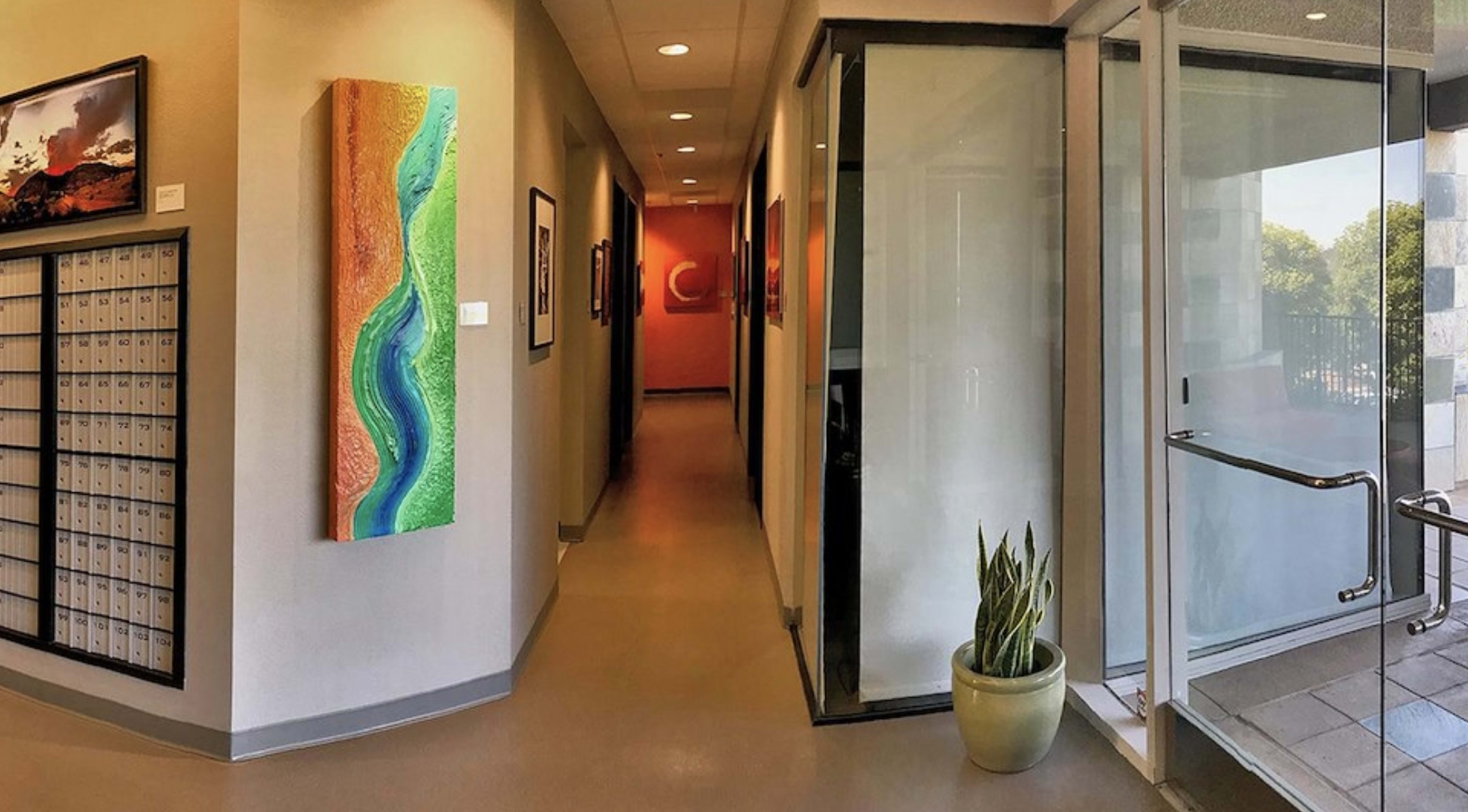 The image shows a hallway with mailboxes on one side, colorful artwork on the wall, and glass walls leading to an outdoor area.