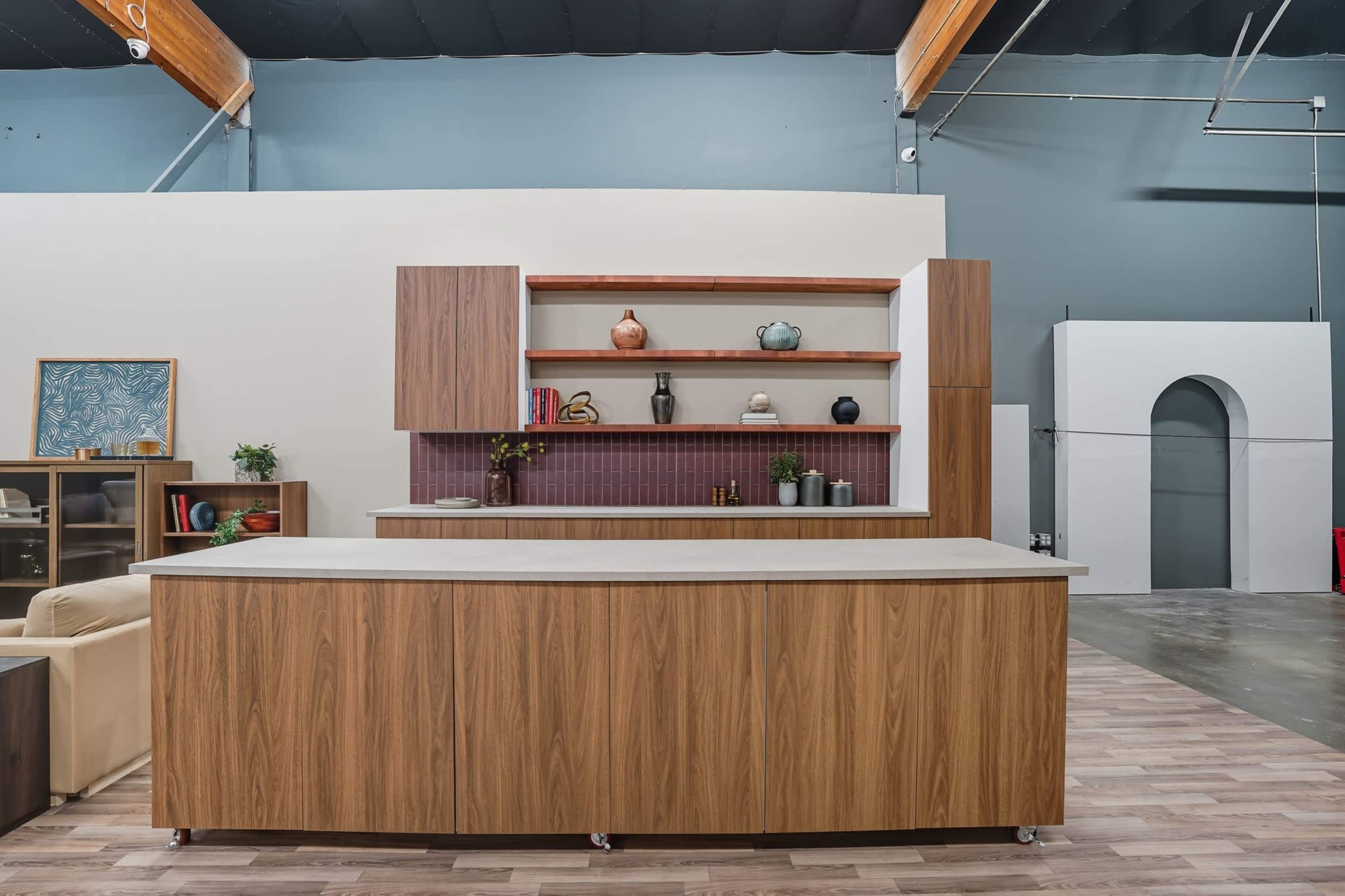 The image shows a modern kitchen space featuring a central wooden island with a gray countertop, wall-mounted shelves with decorative items, and a minimalist design.