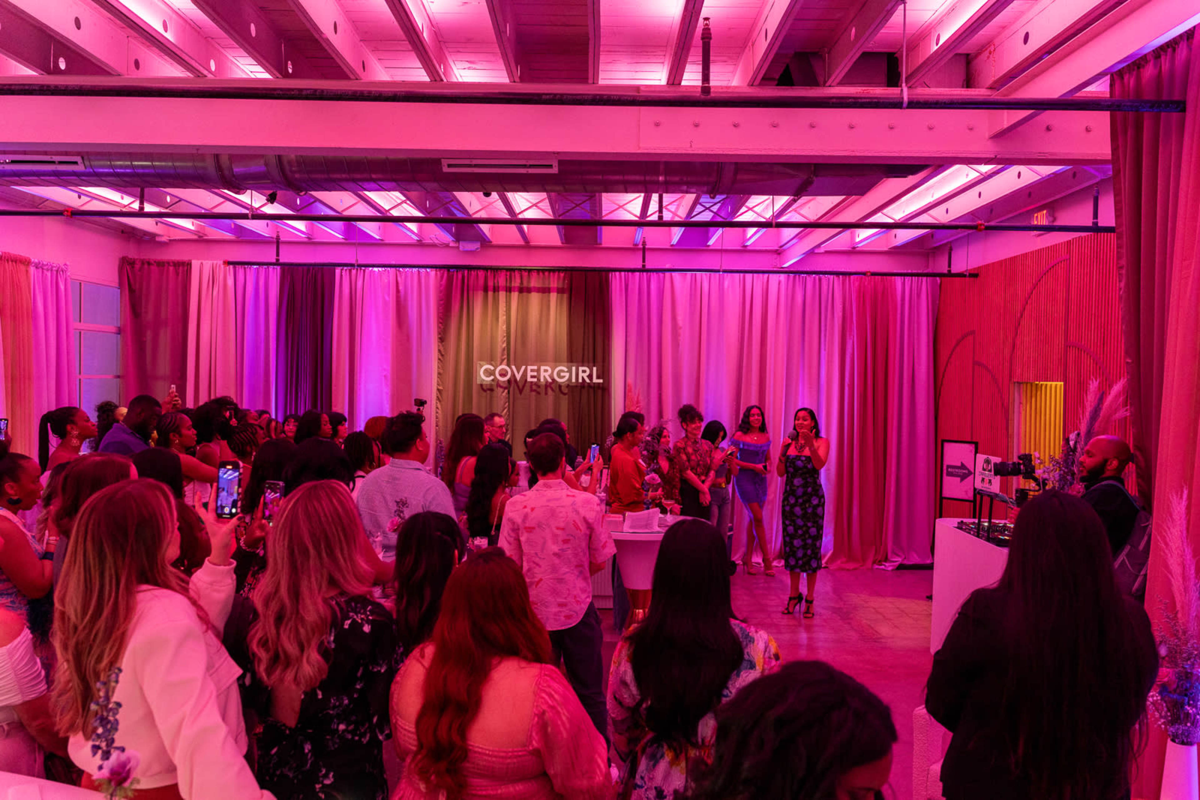 A large group of people gathers in a brightly lit venue with pink lighting as a woman speaks on stage.