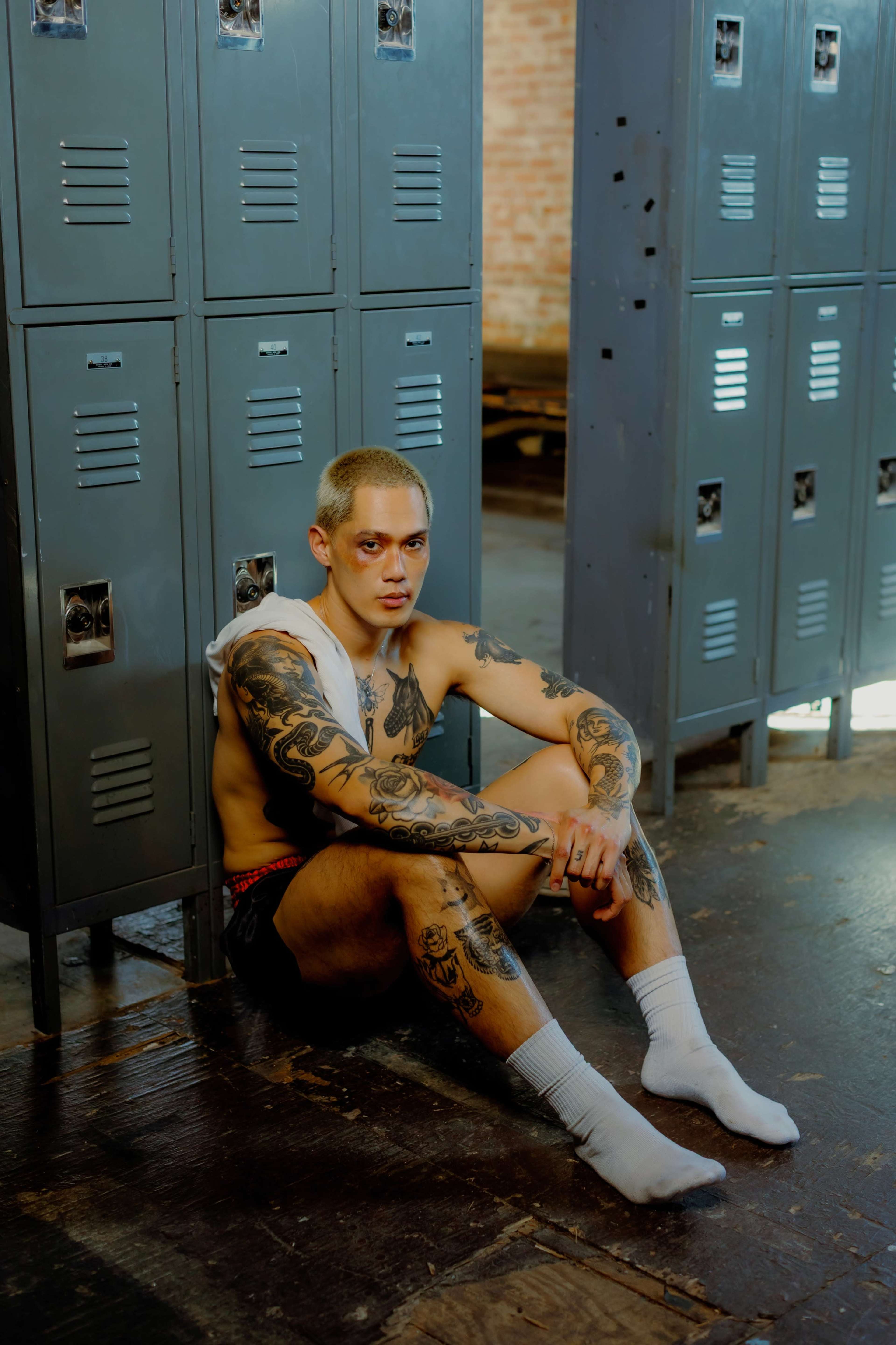 A tattooed person sits on the floor next to lockers in a dimly lit room.