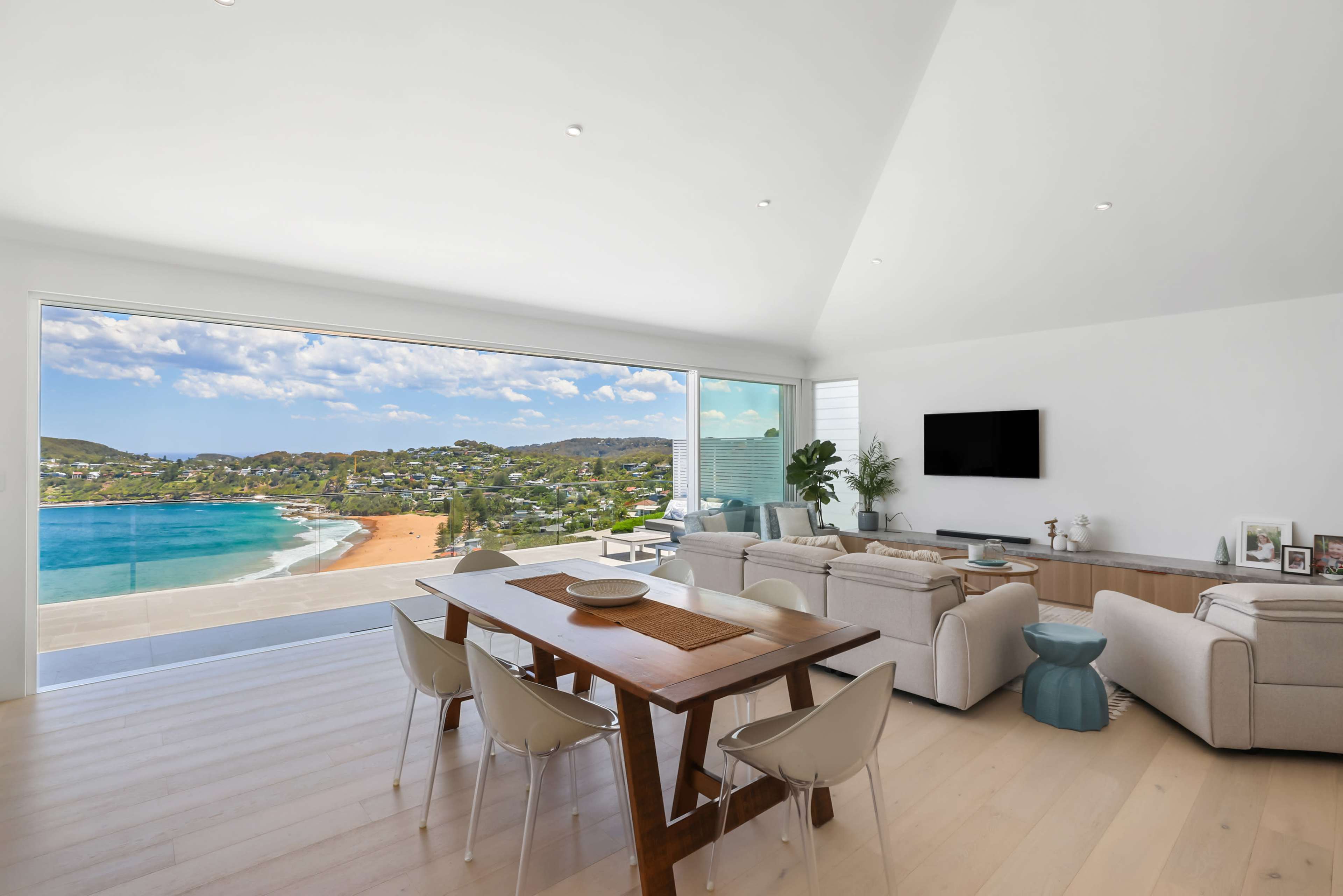 A bright, modern living space features a dining area with a wooden table, surrounded by light chairs, and offers a panoramic view of the beach and coastline through large glass doors.