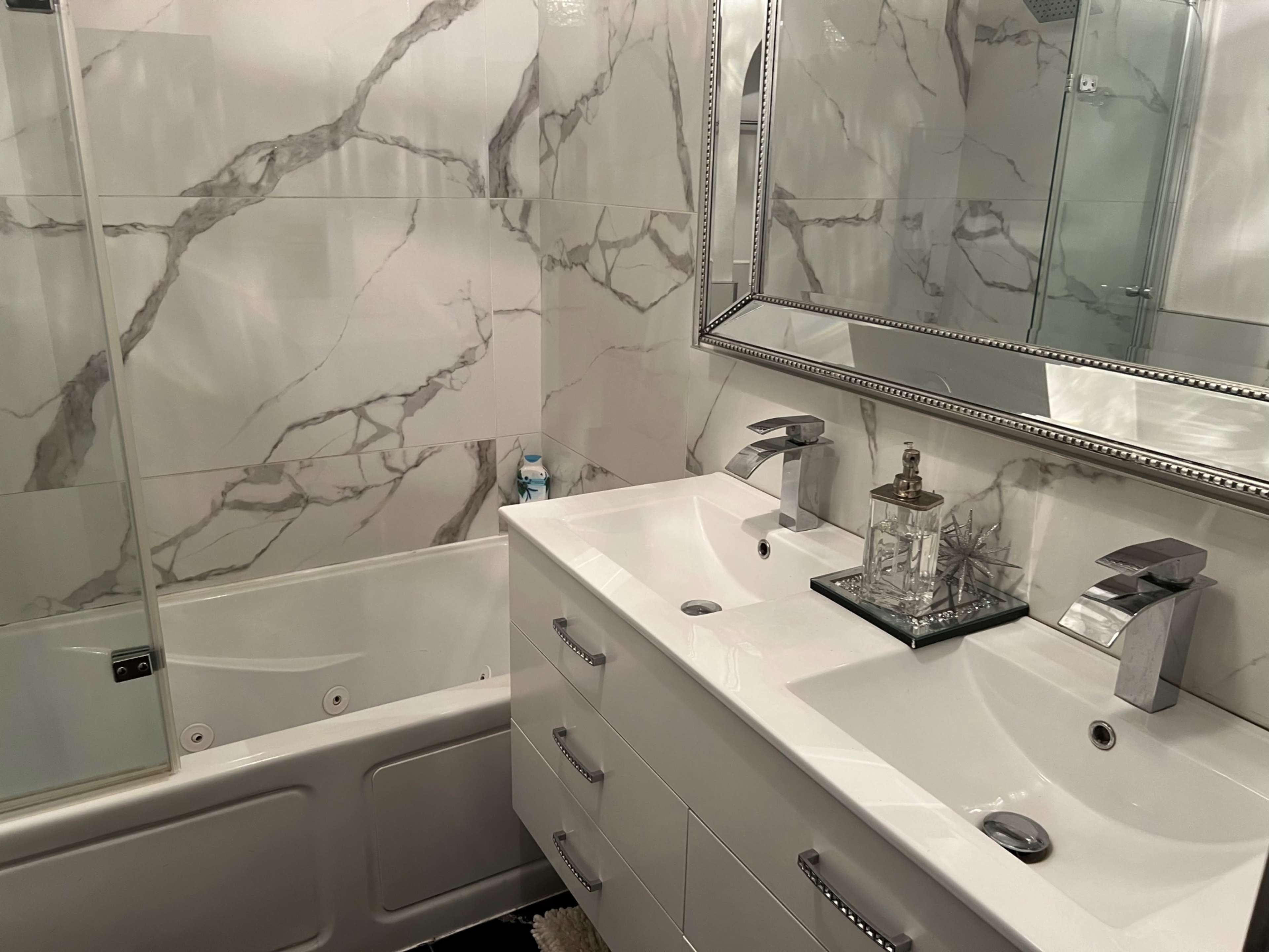 The image shows a modern bathroom featuring a bathtub, a double sink vanity with sleek faucets, and a large mirror against a marble-patterned wall.