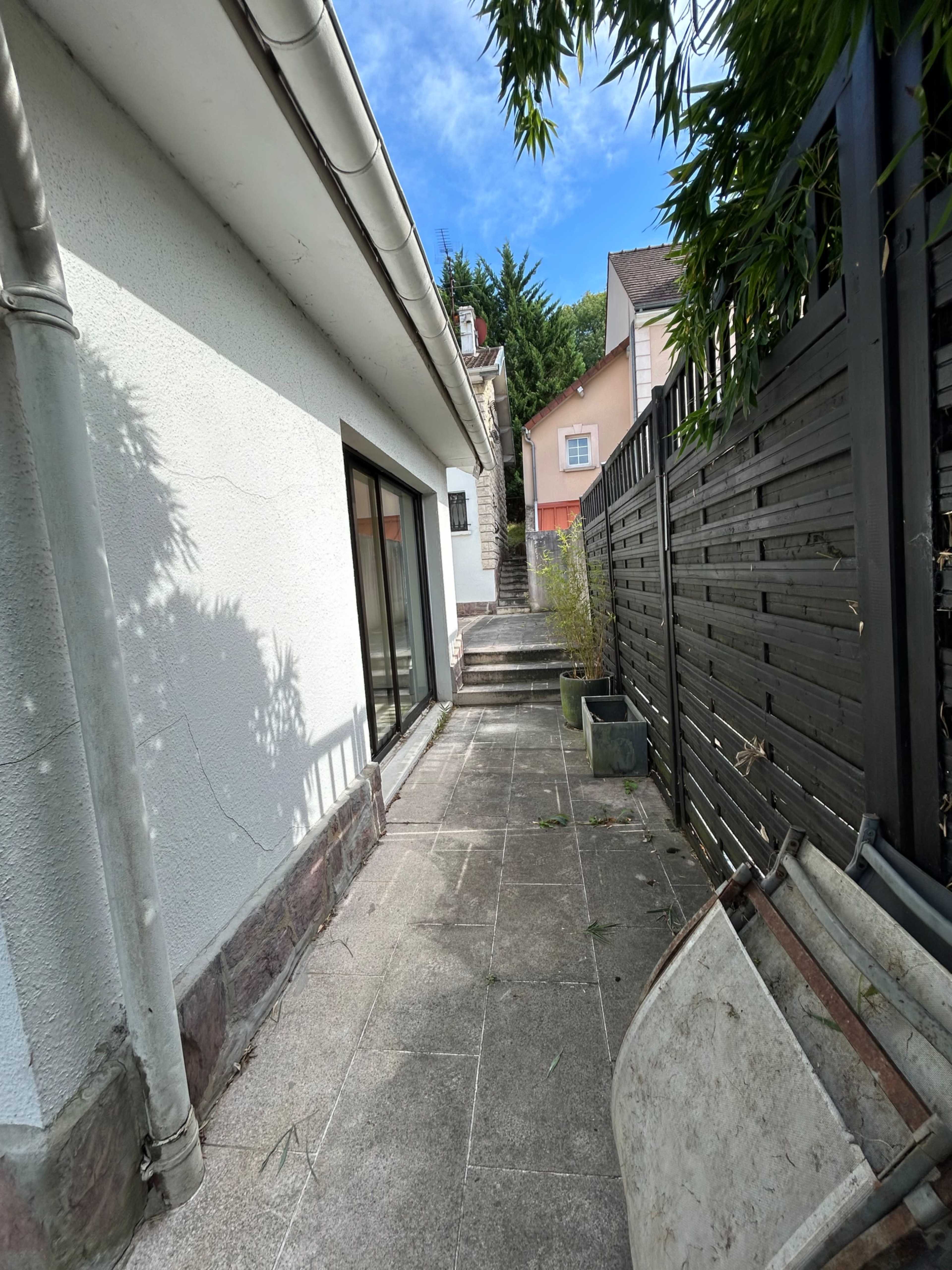 The image shows a narrow walkway beside a house, leading to a staircase, with a wooden fence on one side and a few potted plants.
