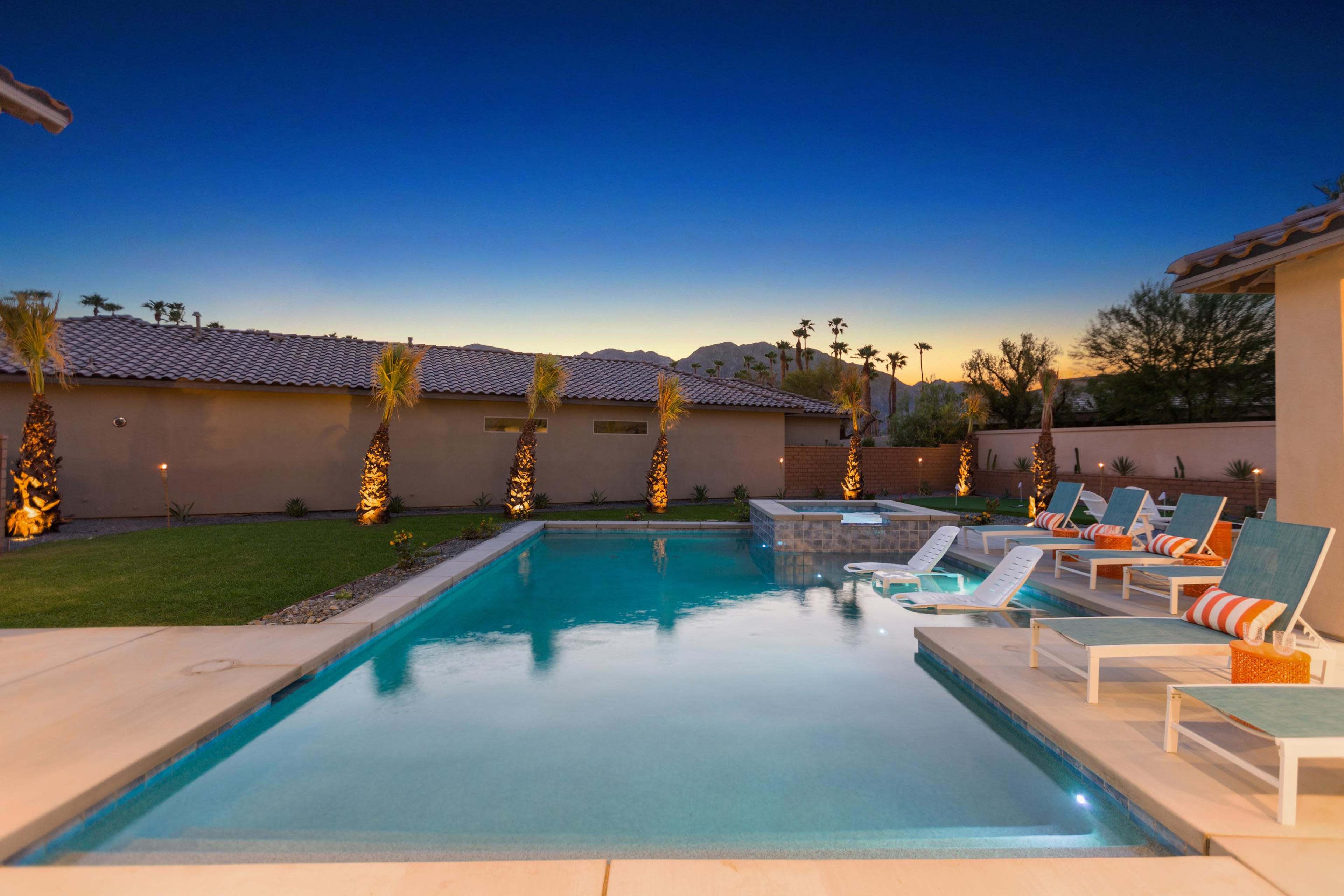 A rectangular swimming pool with lounge chairs surrounds it, accompanied by palm trees and a mountain backdrop at dusk.