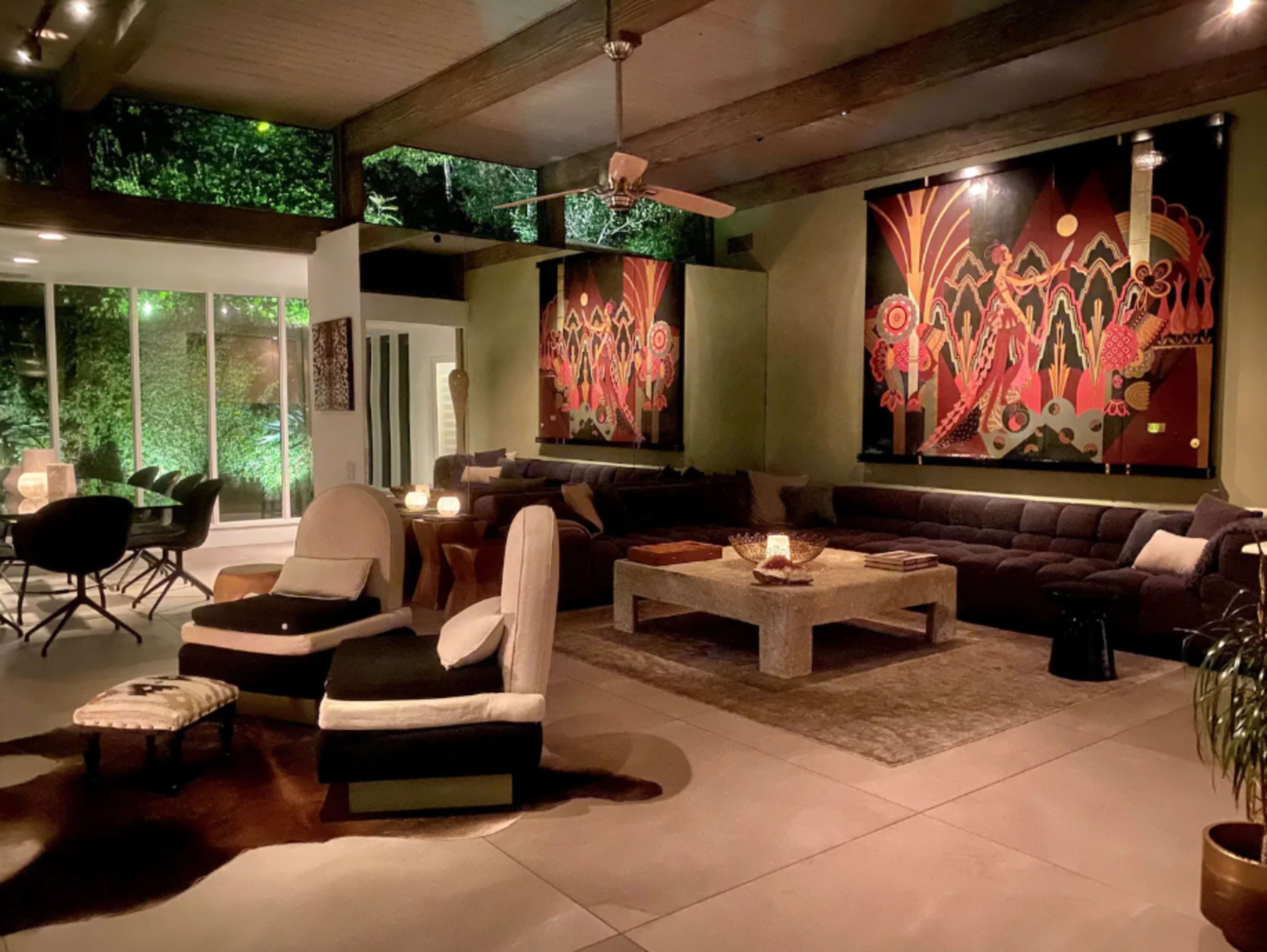 The image shows a modern living room with large windows, a sectional sofa, and colorful wall art, surrounded by greenery.