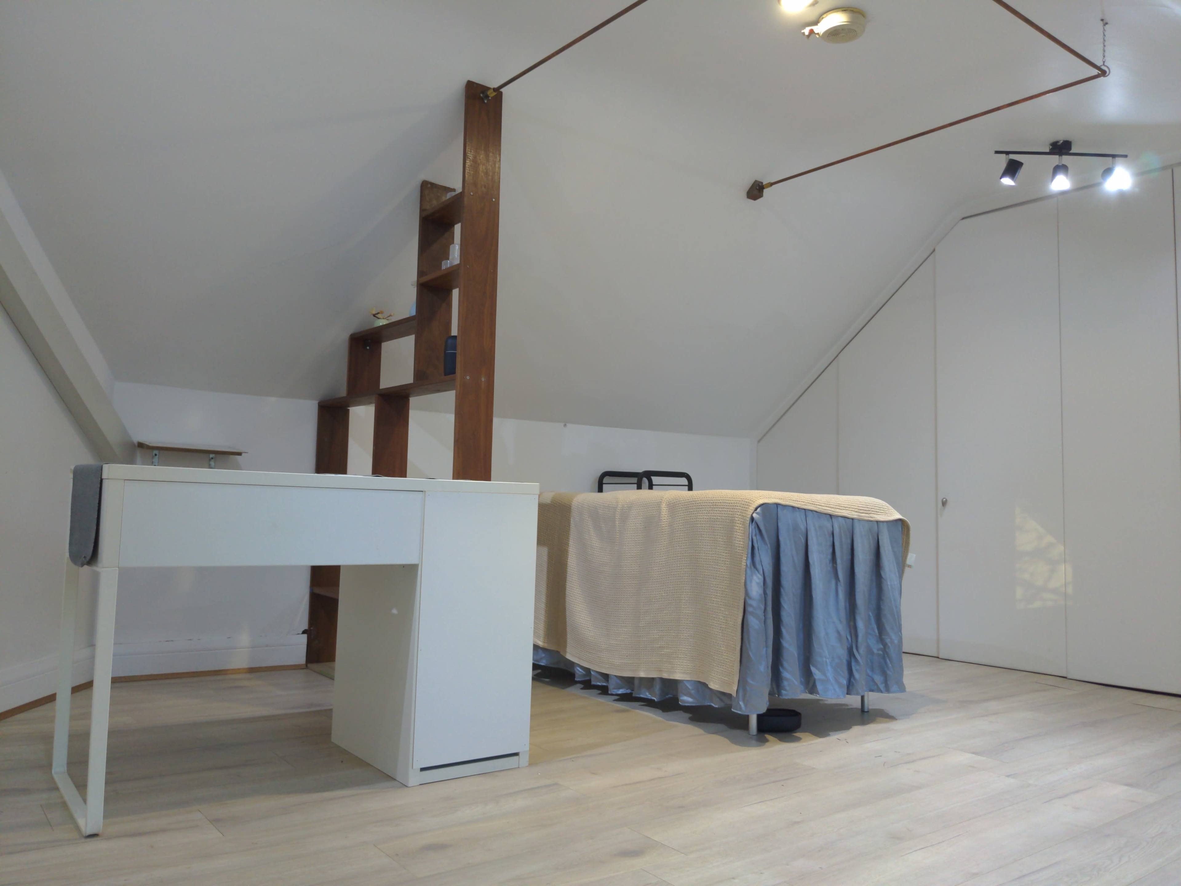 The image shows a sparsely furnished attic room featuring a bed with a light-colored bedspread, a desk, and a shelving unit against one wall.