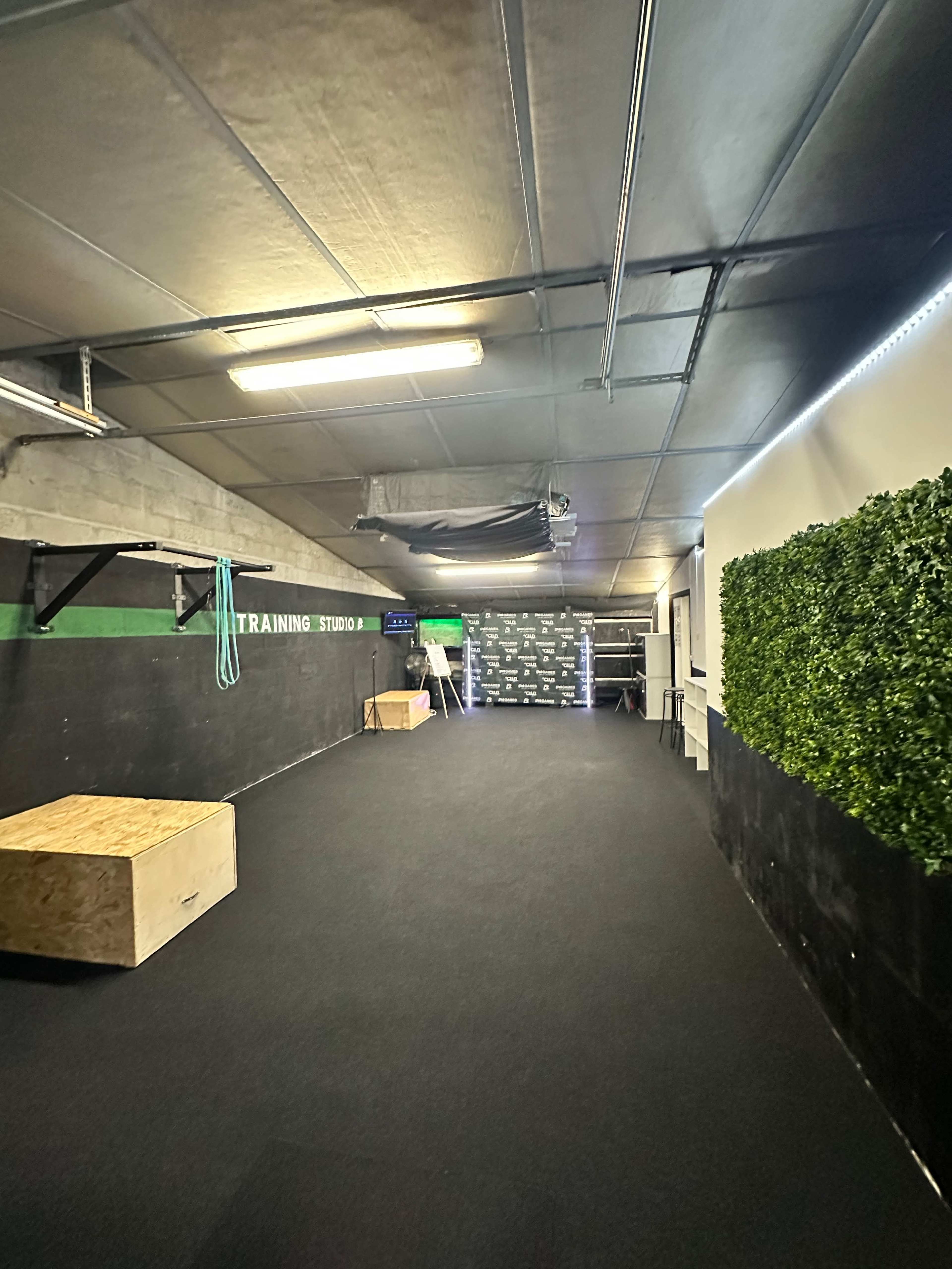 The image shows an indoor training studio with a black rubber floor, wooden boxes for exercises, and green wall accents, along with a television mounted on a wall.