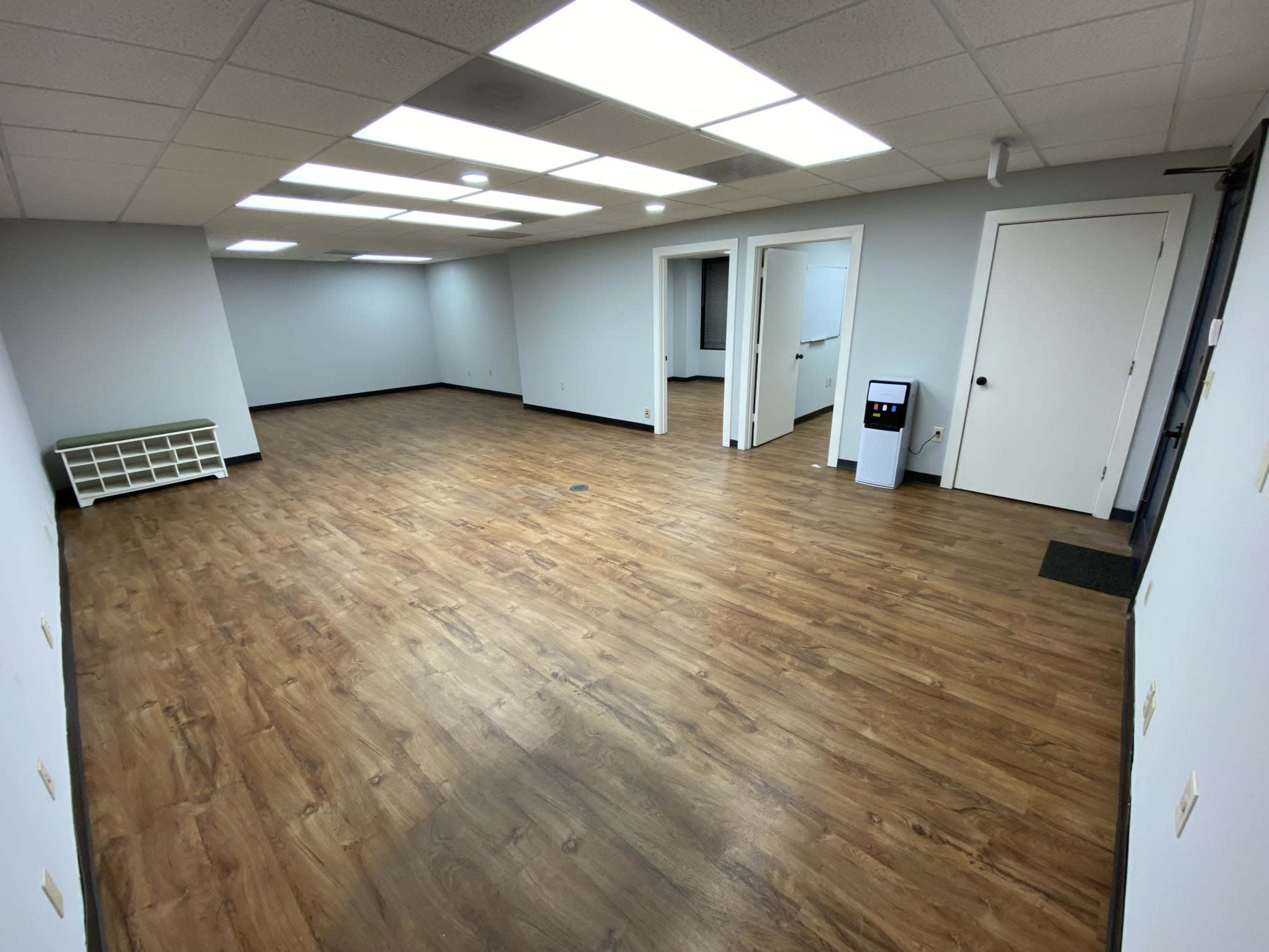 The image shows an empty office space with wooden laminate flooring and gray walls, illuminated by overhead fluorescent lights.