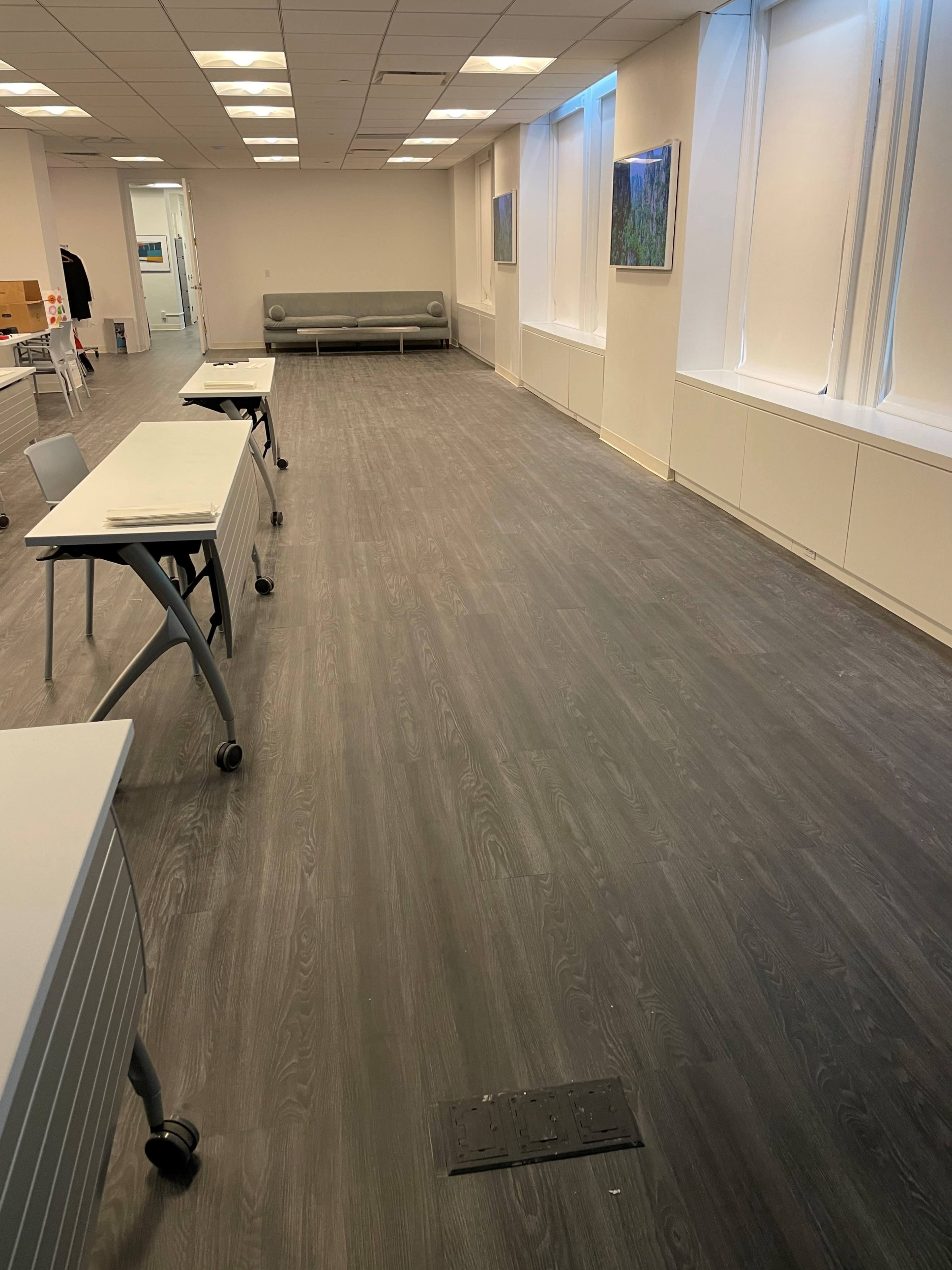 A large, empty room with gray flooring, white walls, and several tables arranged in a row, along with a couch and artwork on the walls.