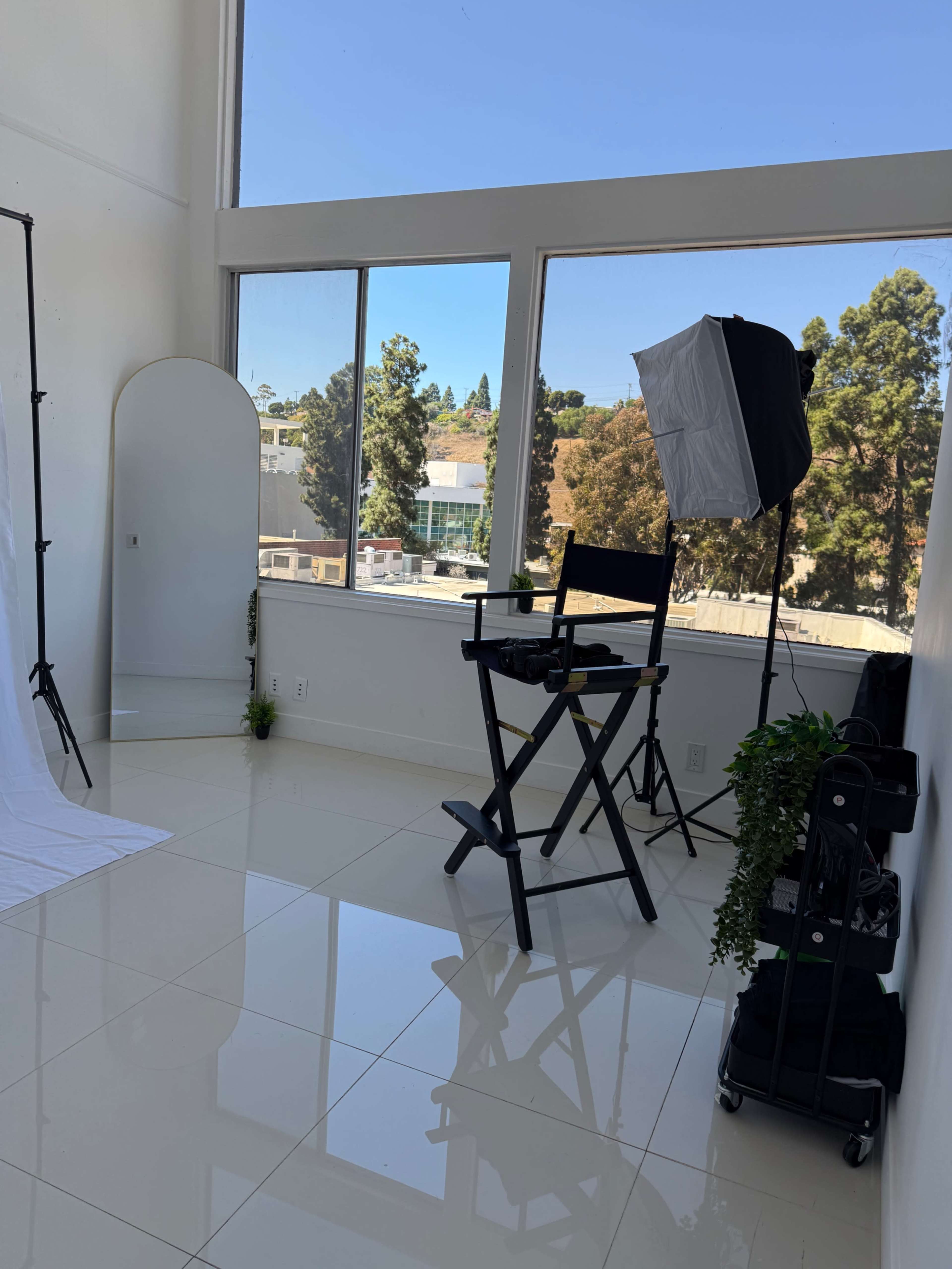 The image shows a bright photo studio with large windows, featuring a director's chair, a lighting setup, a mirror, and greenery in the corners.