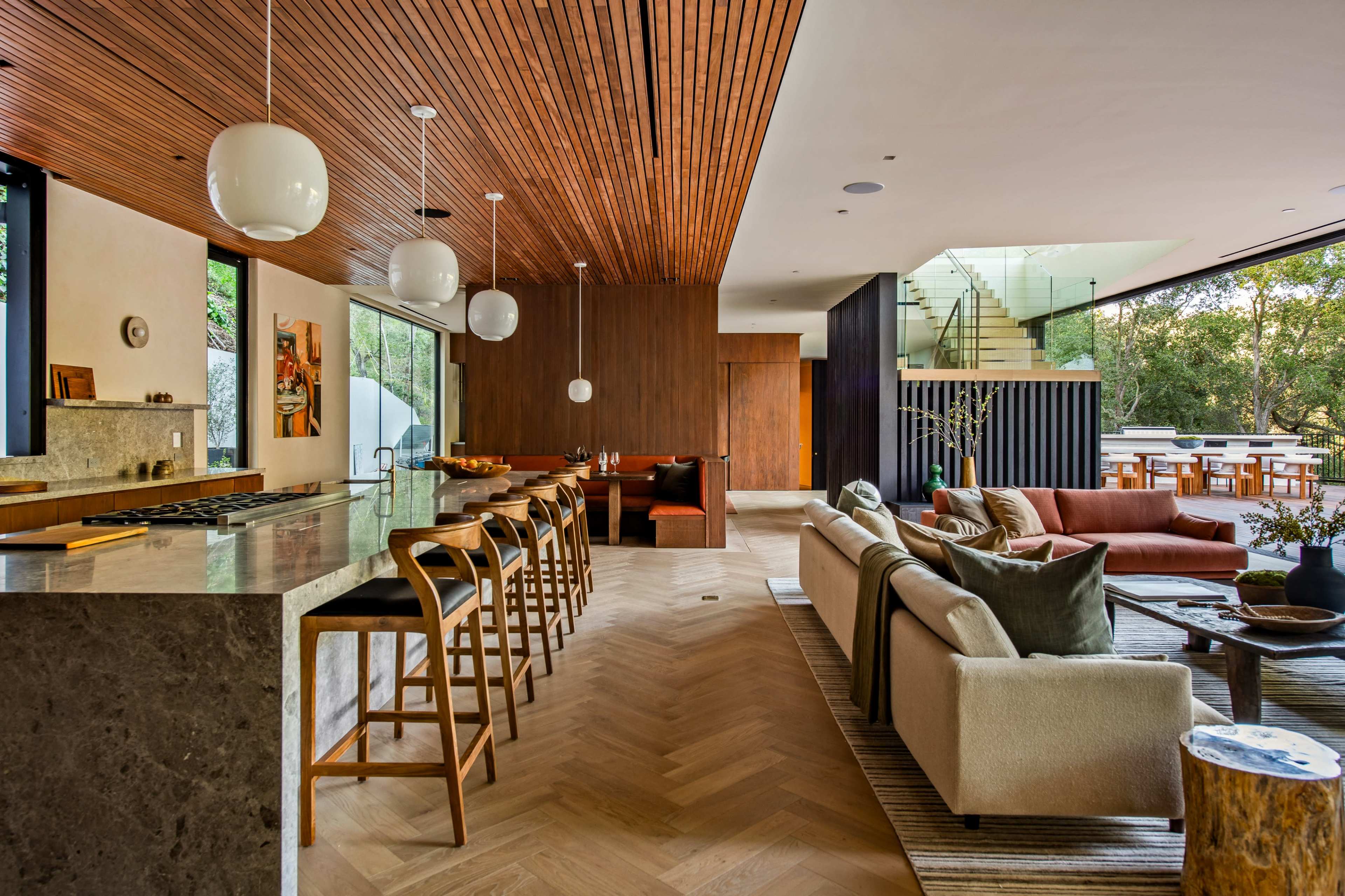 The image shows a modern open-concept living and dining area with wooden ceilings, a kitchen island, and large windows that provide natural light.