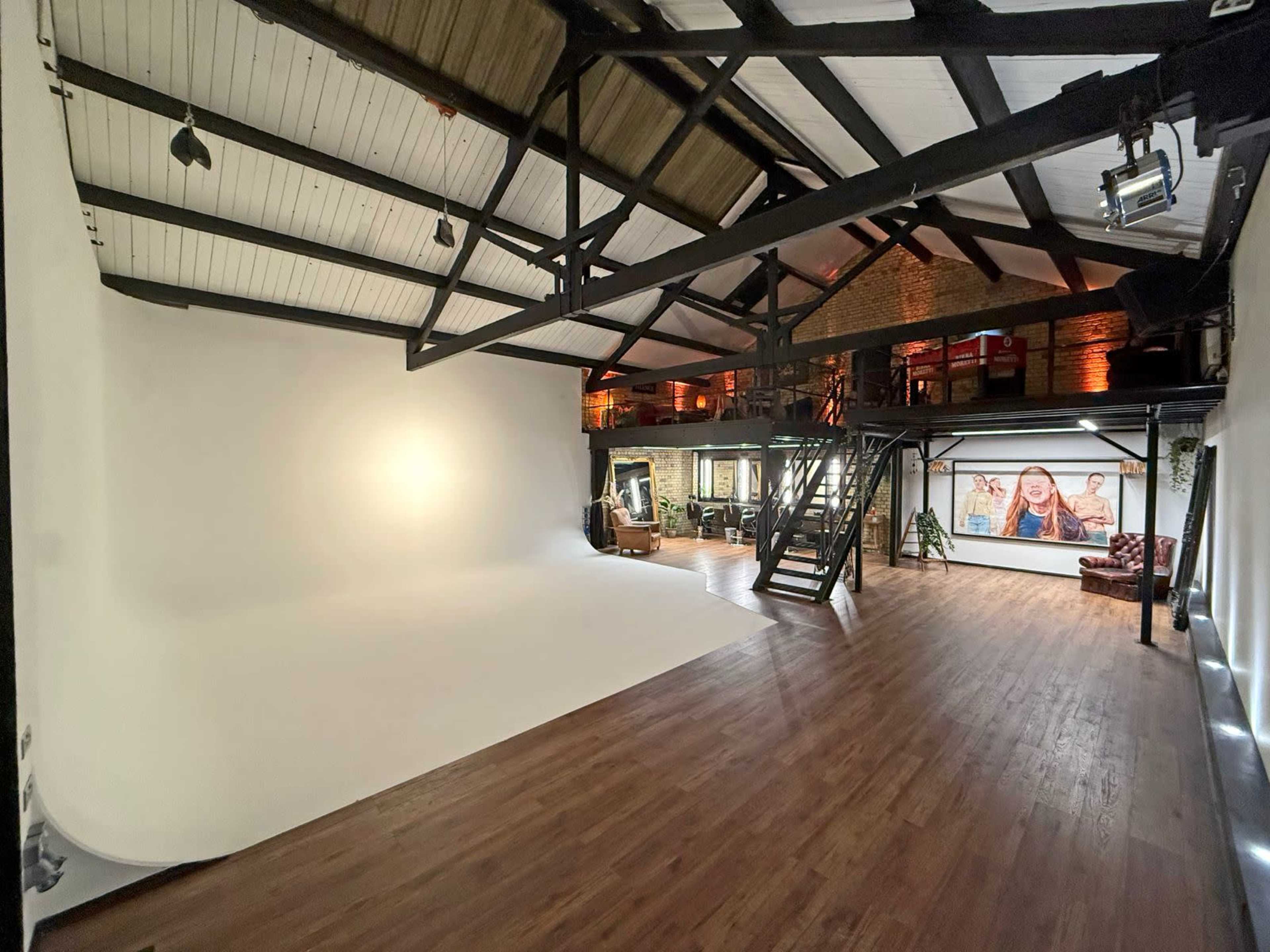 The image shows a spacious photography studio with a large white backdrop, wooden floors, and exposed beams, along with a staircase and an area featuring a large portrait on the wall.