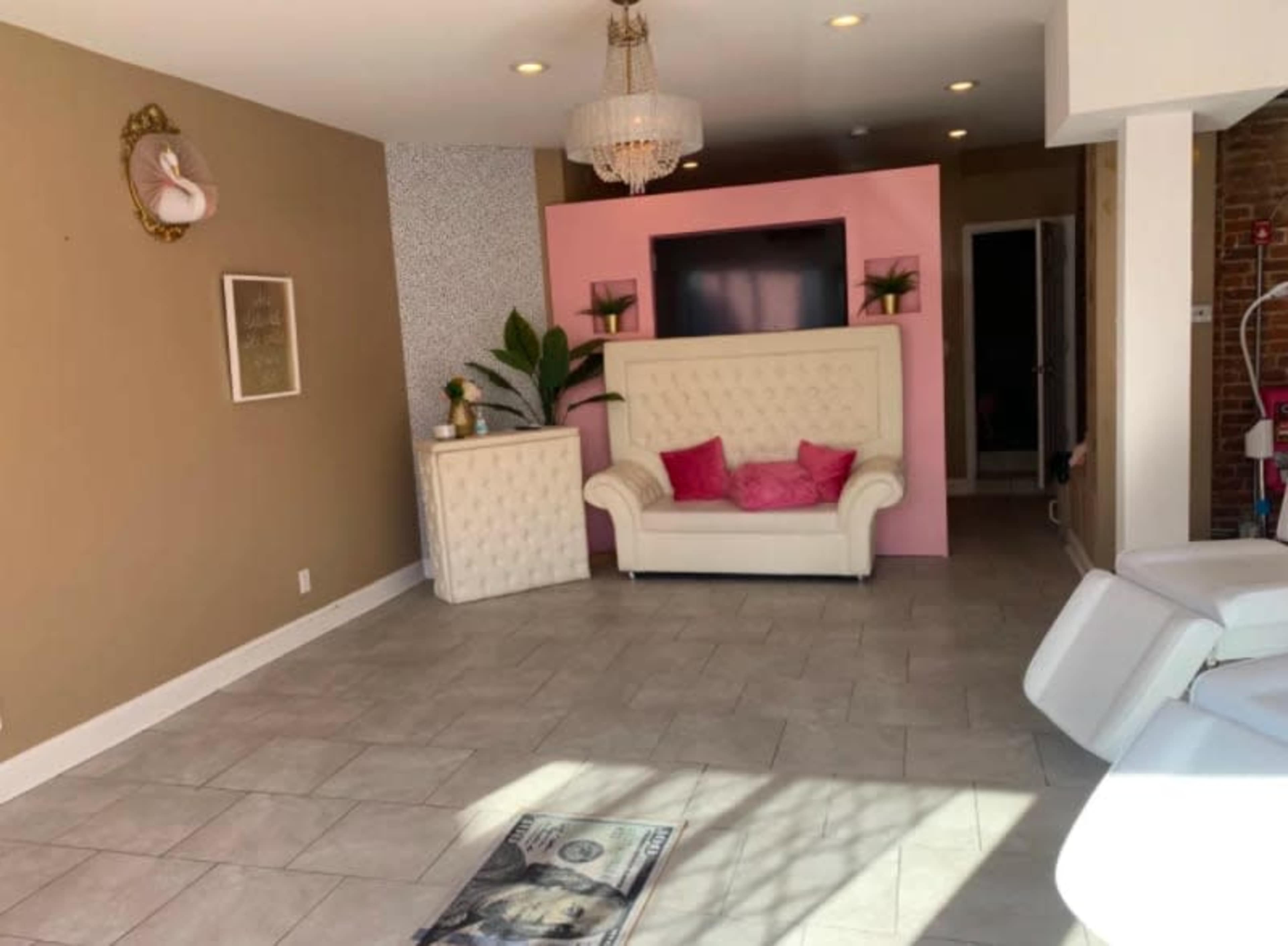 A spacious, brightly lit room with light tile flooring, featuring a white couch with pink accents, a wall-mounted TV, and decorative plants.