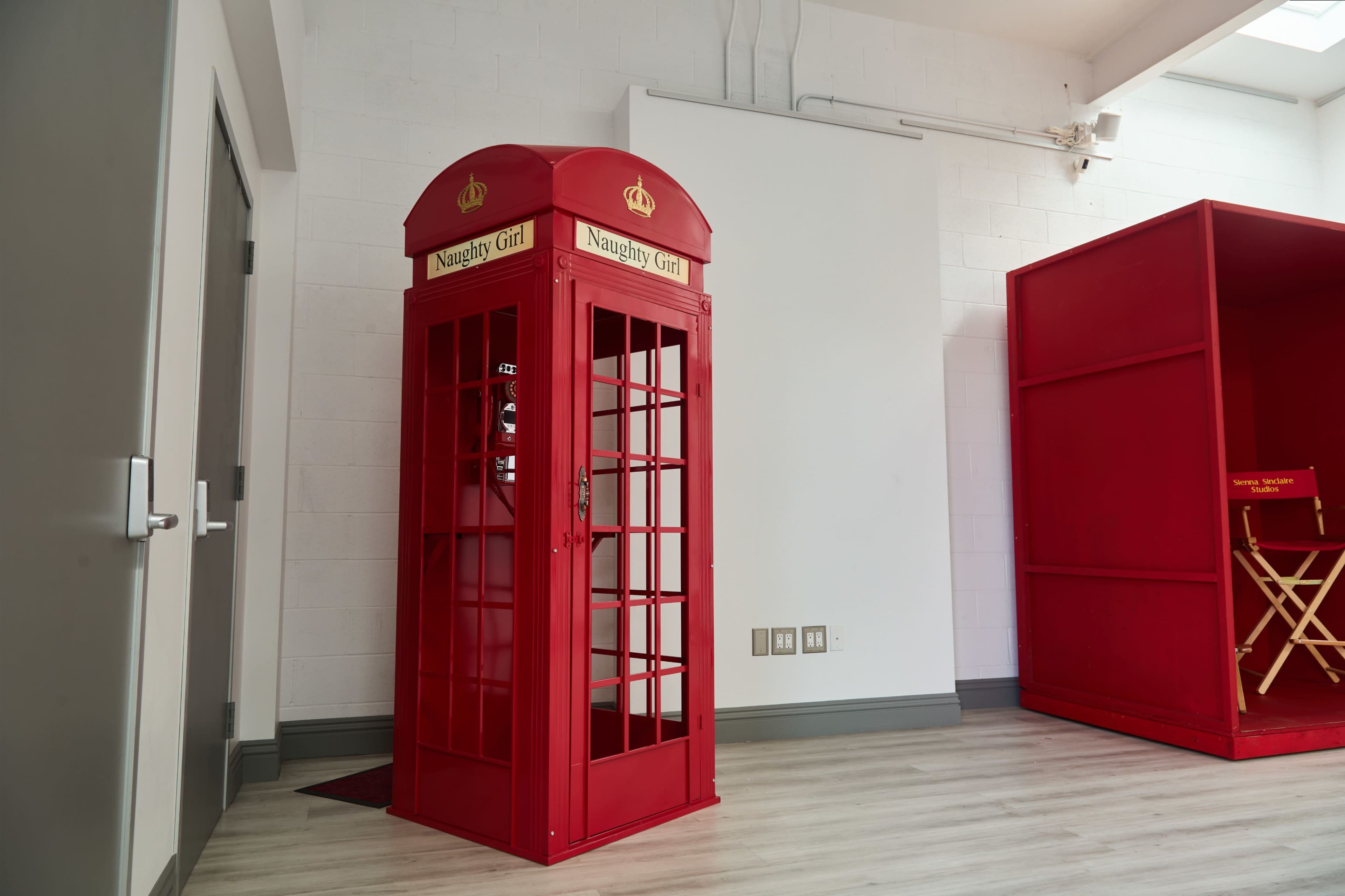 A red telephone booth labeled "Naughty Girl" stands in a well-lit room with white walls and light wooden flooring.