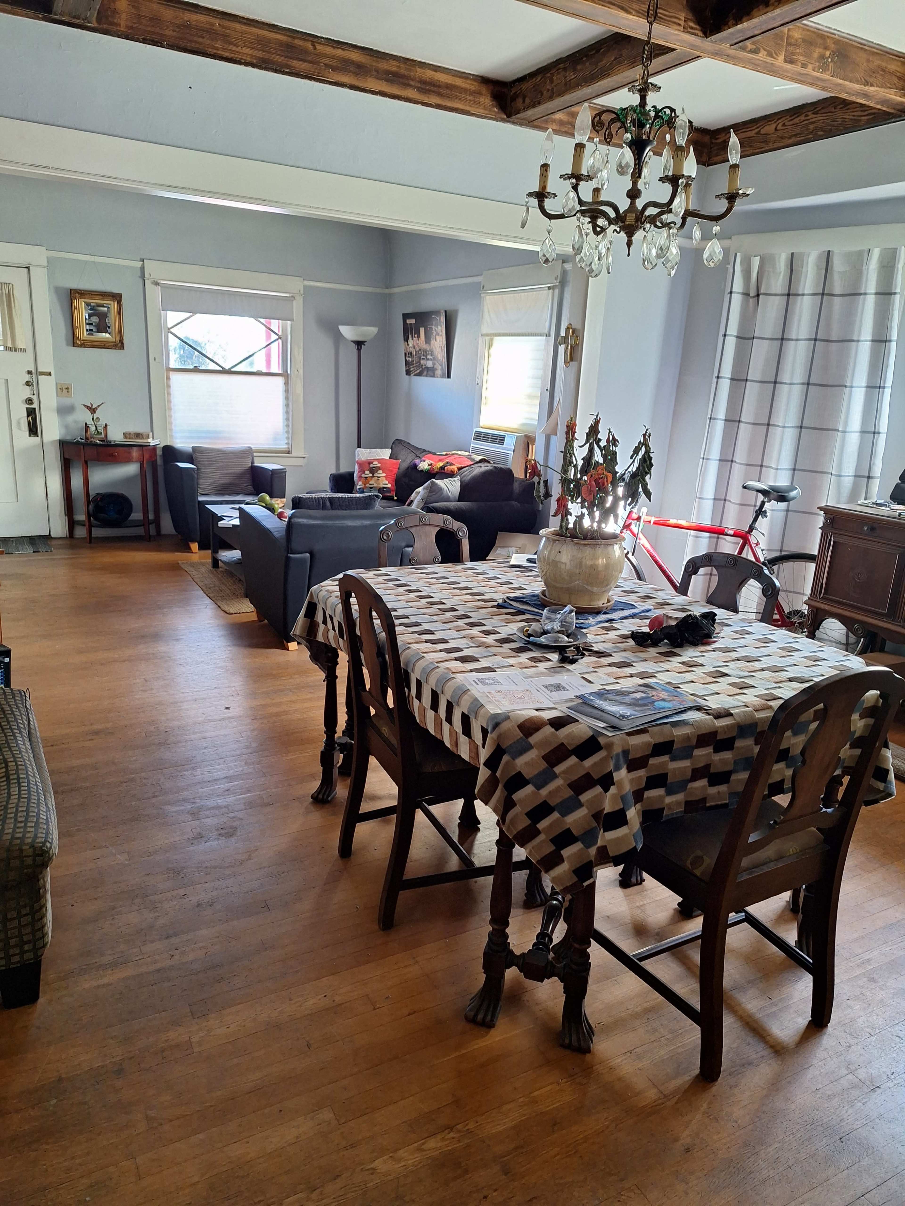 The image shows a spacious room with a dining table at the center, surrounded by chairs, and a living area with a sofa and bicycle visible in the background.