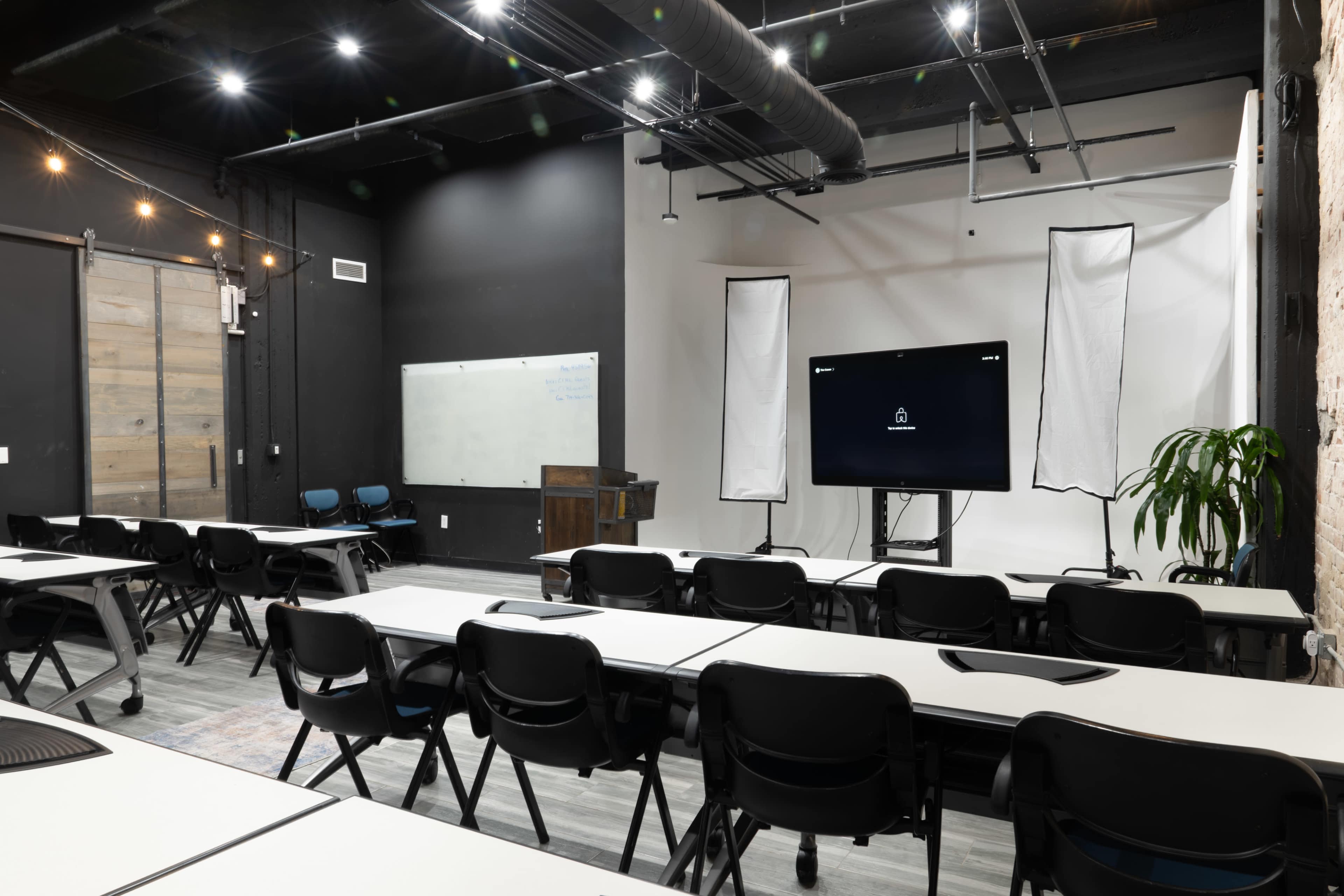 The image shows a modern classroom setting with rows of black chairs and white tables facing a large display screen, flanked by two backdrops, in a well-lit, industrial-style room.
