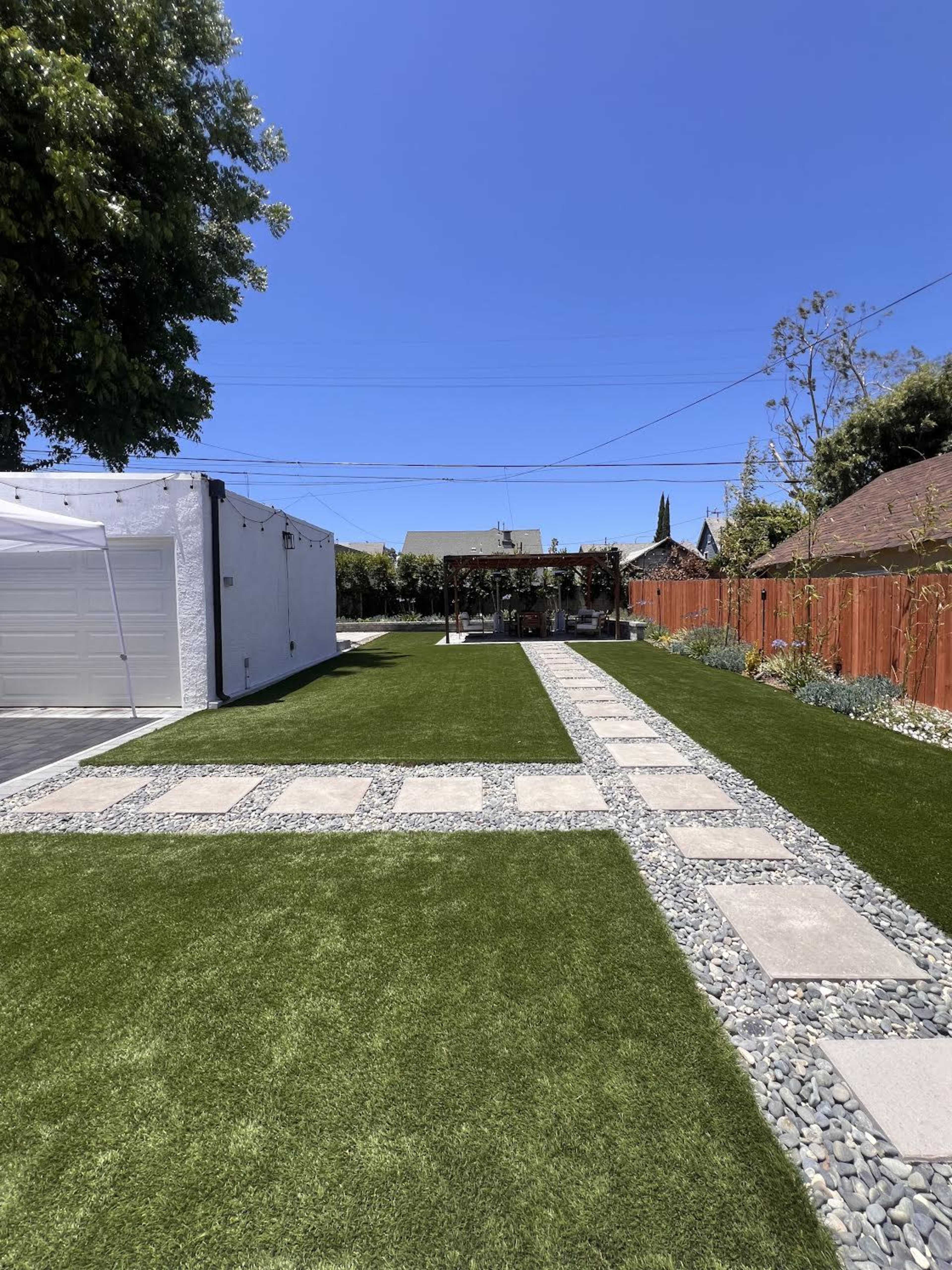 The image shows a landscaped backyard featuring a pathway of pavers and gravel leading to a pergola, surrounded by green artificial grass and wooden fencing.