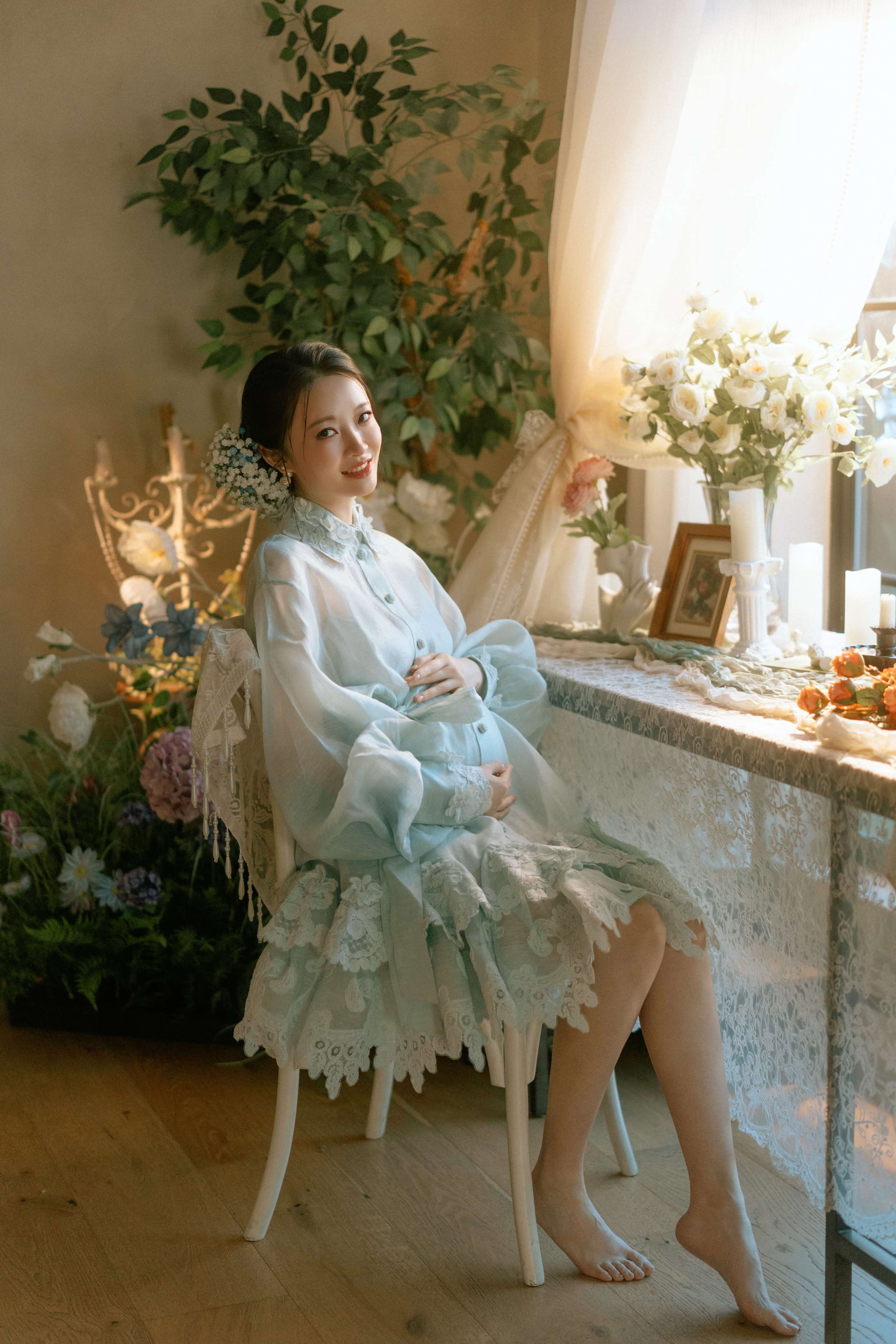 A pregnant woman in a light blue dress sits on a chair beside a table adorned with flowers and photographs in a softly lit room.