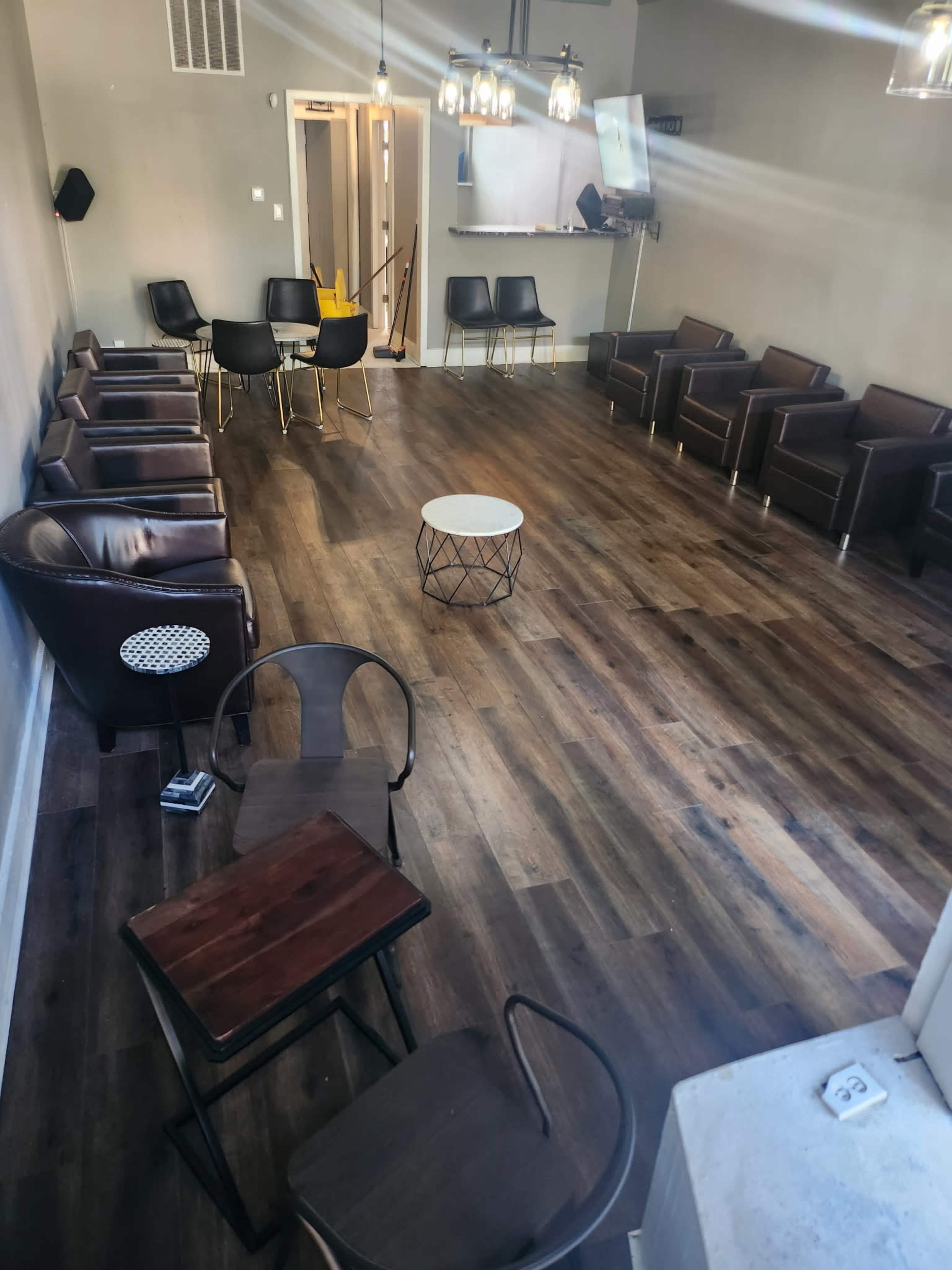 The image shows a spacious, empty room with wooden floors, featuring several brown leather chairs, black dining chairs, and a small white coffee table in the center.