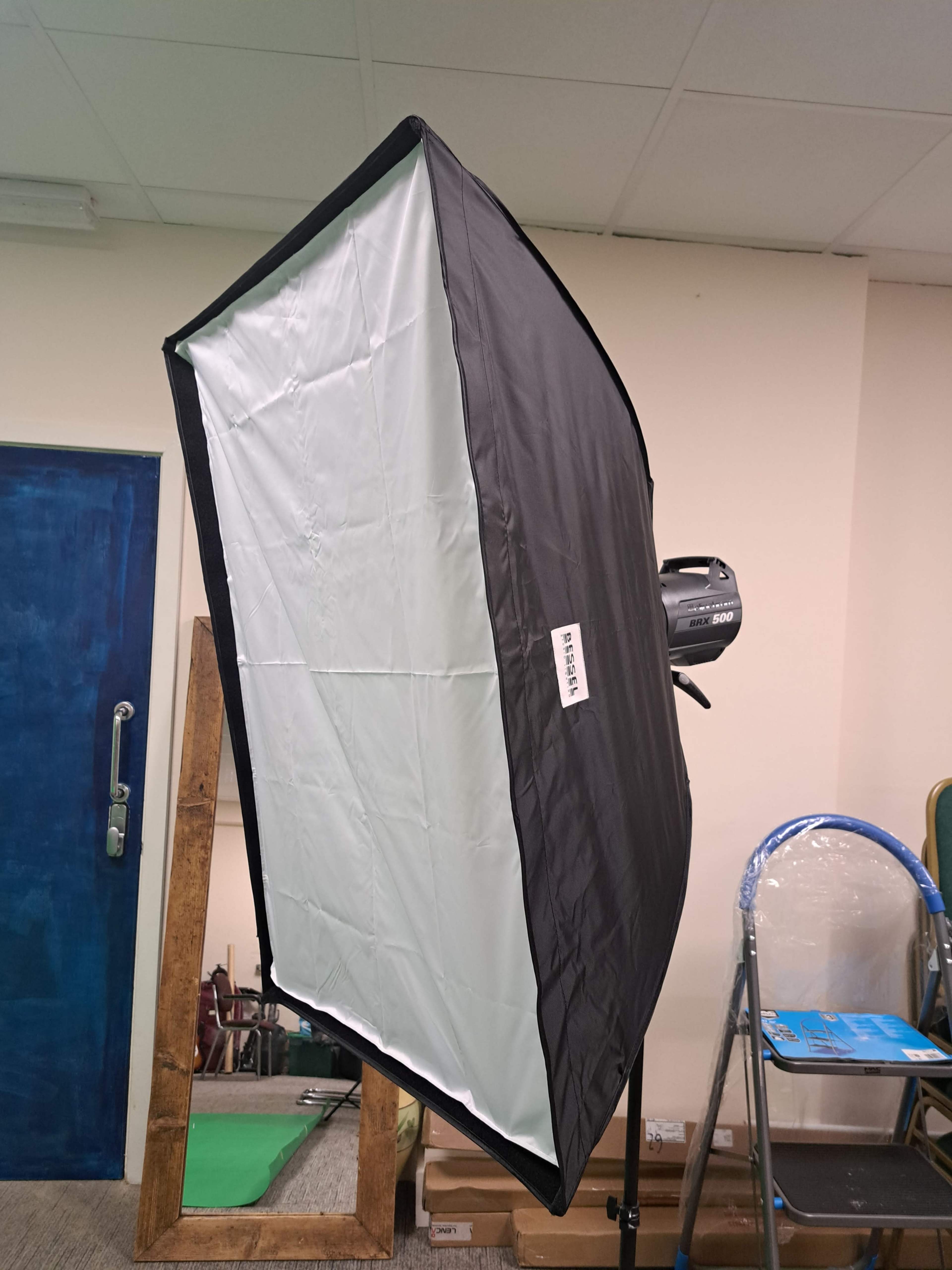 A black softbox is positioned on a light stand inside a room with various equipment in the background.
