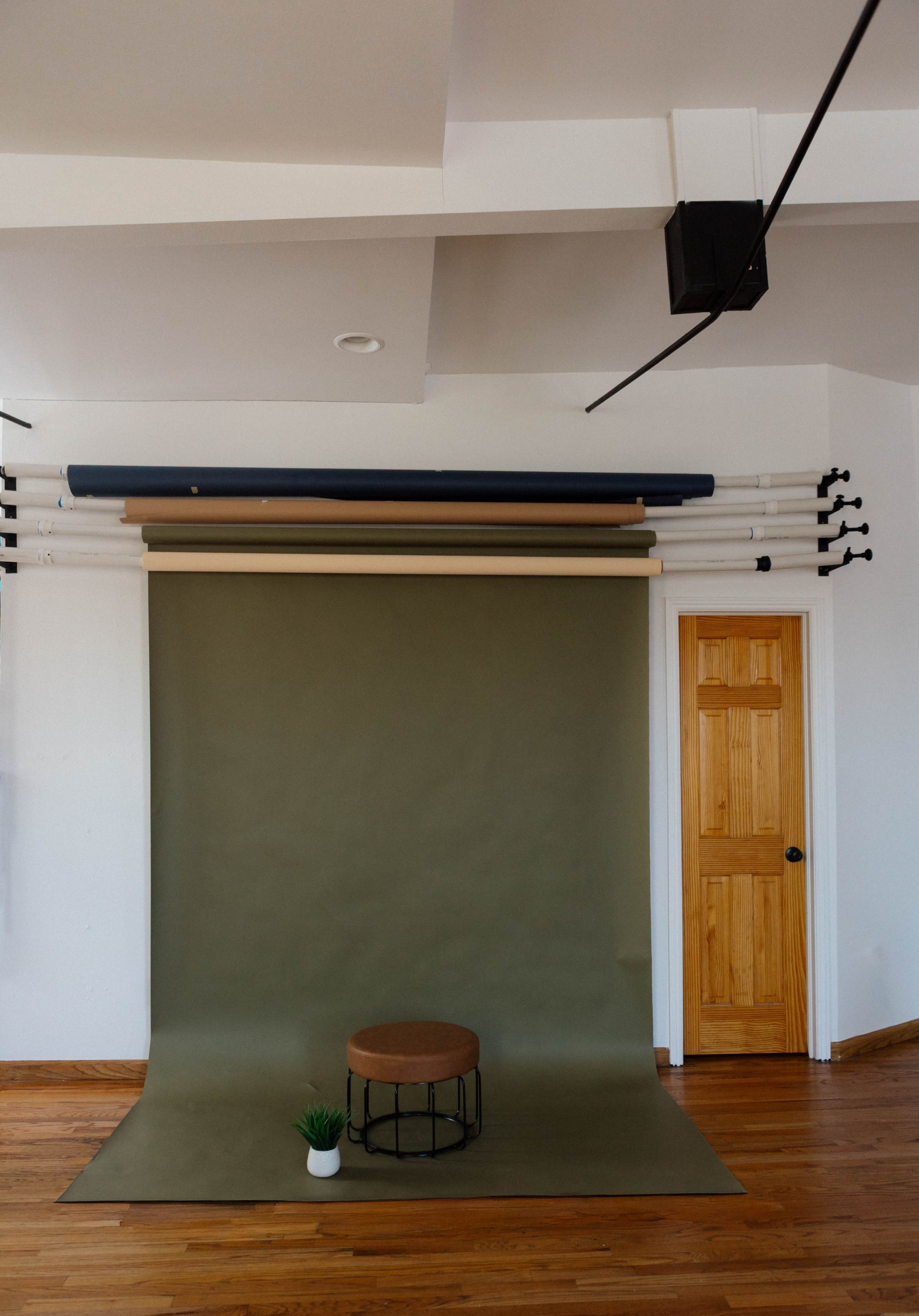 The image shows a photography backdrop setup with a green paper roll, a round stool in front, and wooden flooring in a room with a door.