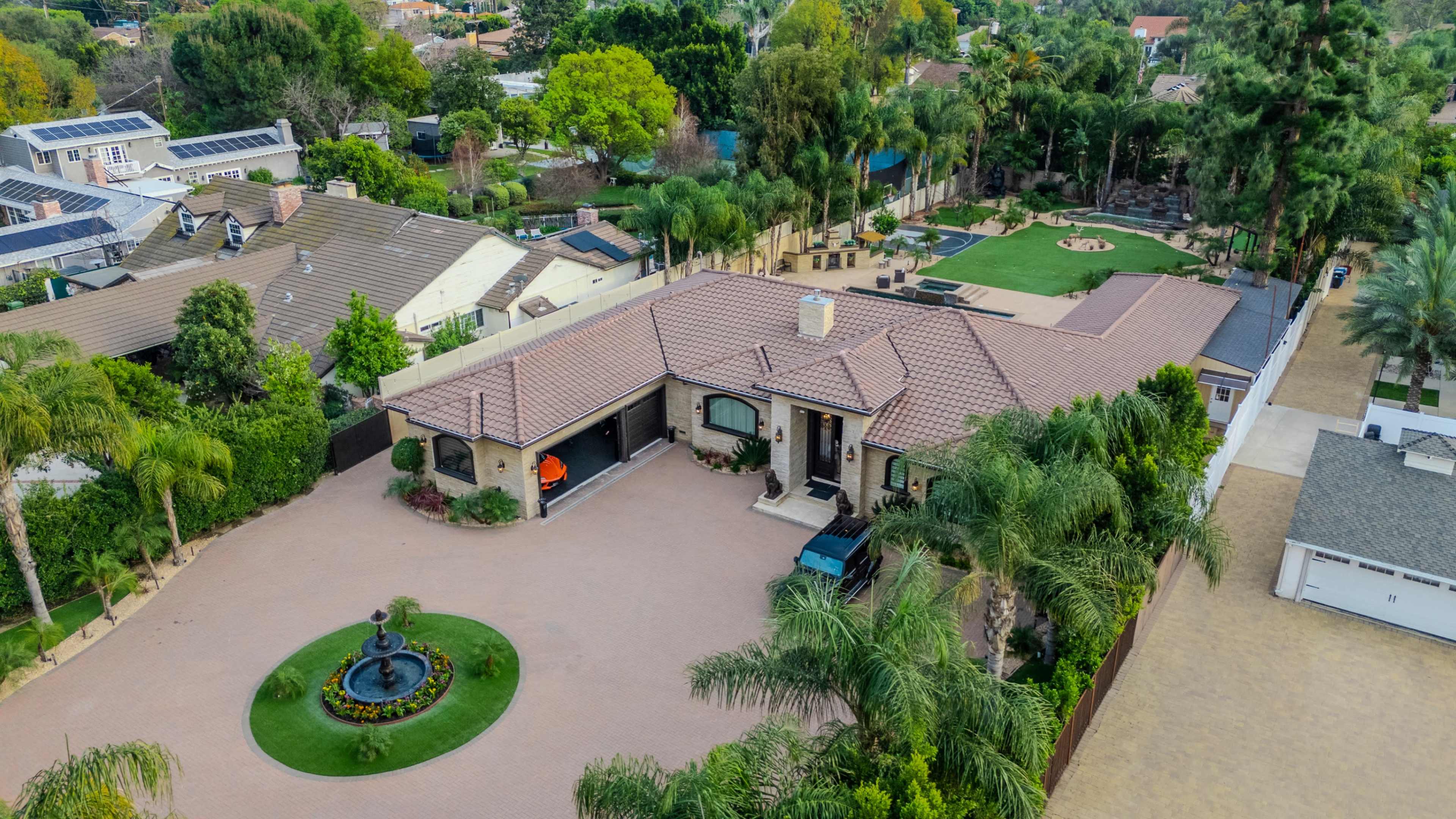 The image shows a large residential property with a landscaped yard, a fountain in front, and neighboring homes in a suburban area.