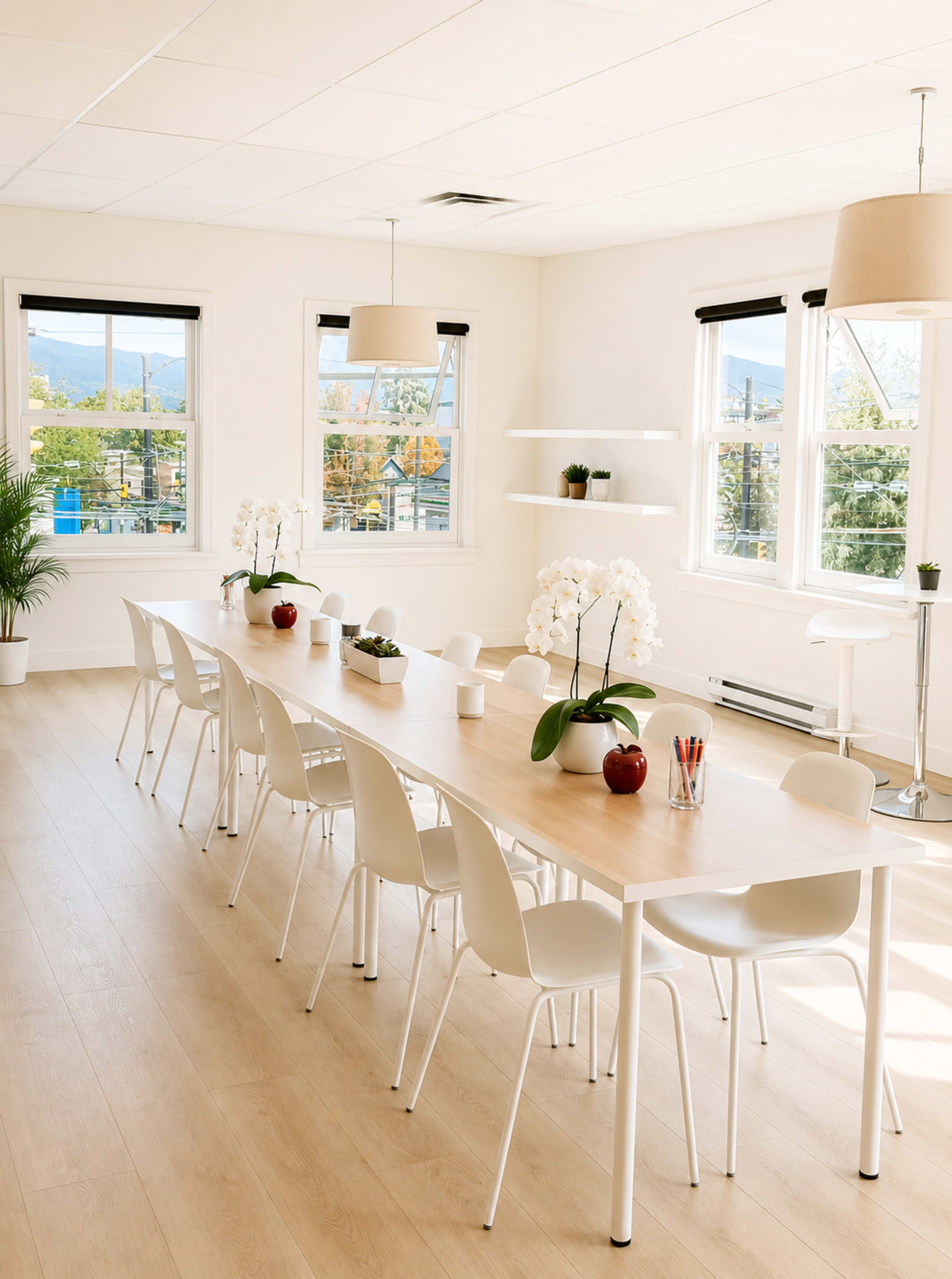 A spacious meeting room features a long wooden table surrounded by white chairs, with large windows allowing natural light and showcasing a view of the outdoors.