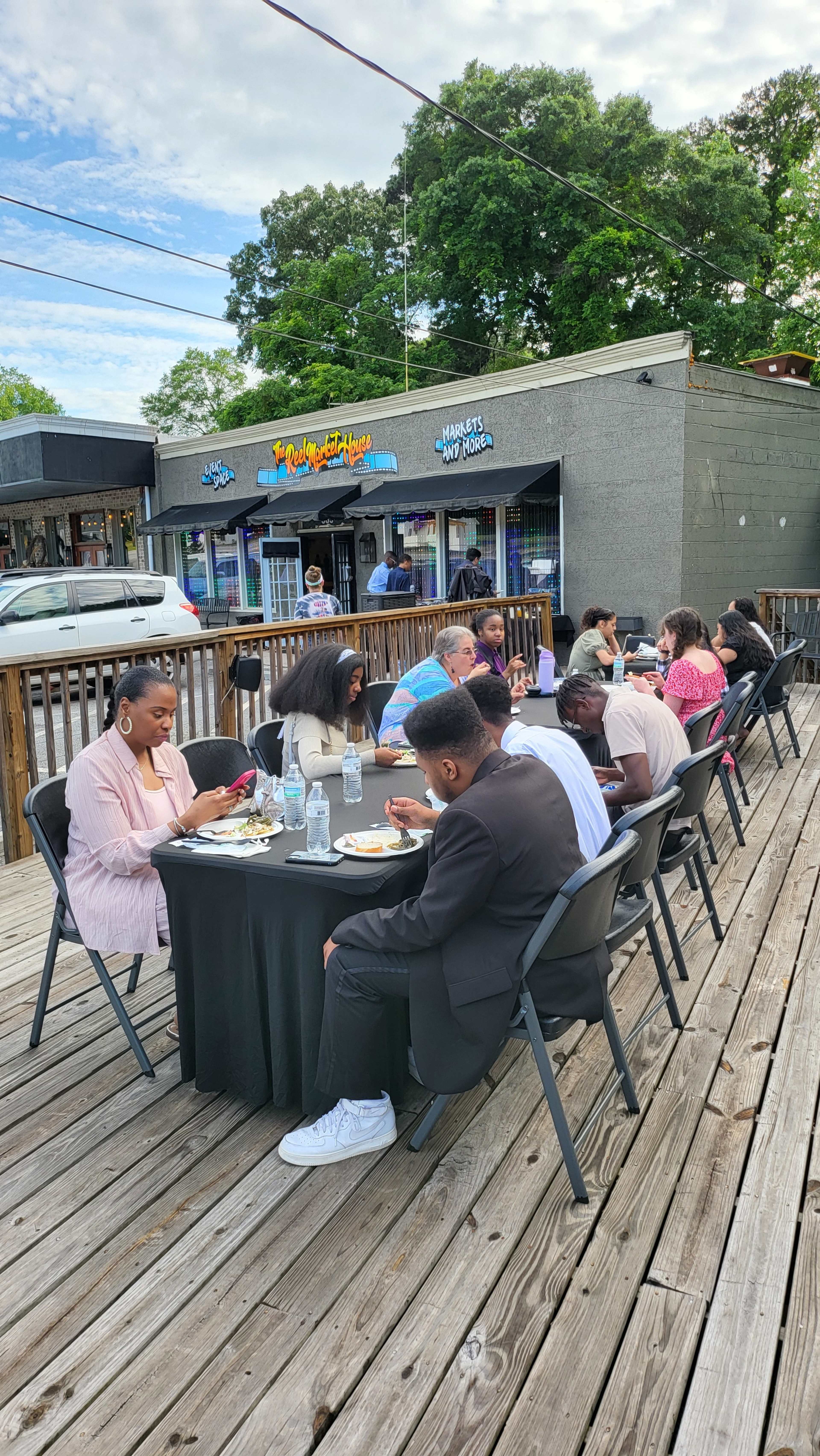 A group of people is seated at tables on a wooden deck outside a restaurant, enjoying meals under a partly cloudy sky.